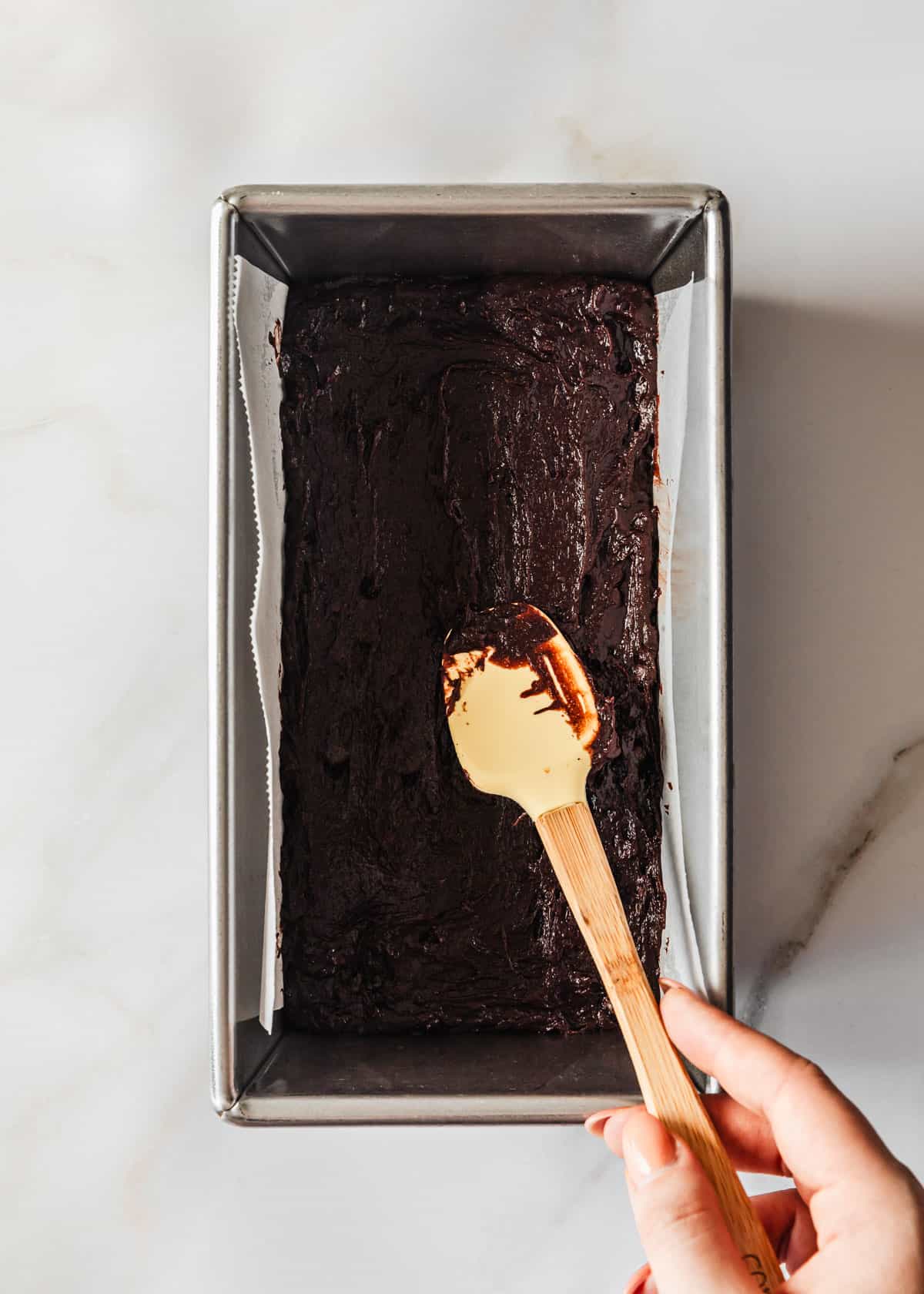 A hand using a rubber spatula to smooth small batch brownies batter in a loaf pan.