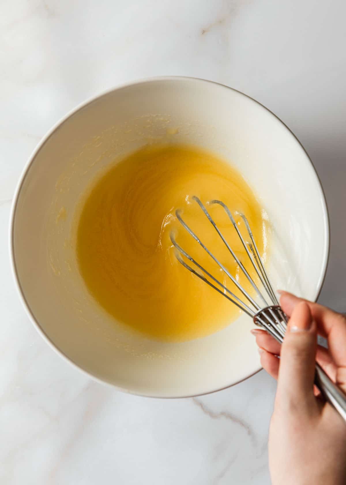 A hand whisking butter and sugar in a white bowl on a white counter.