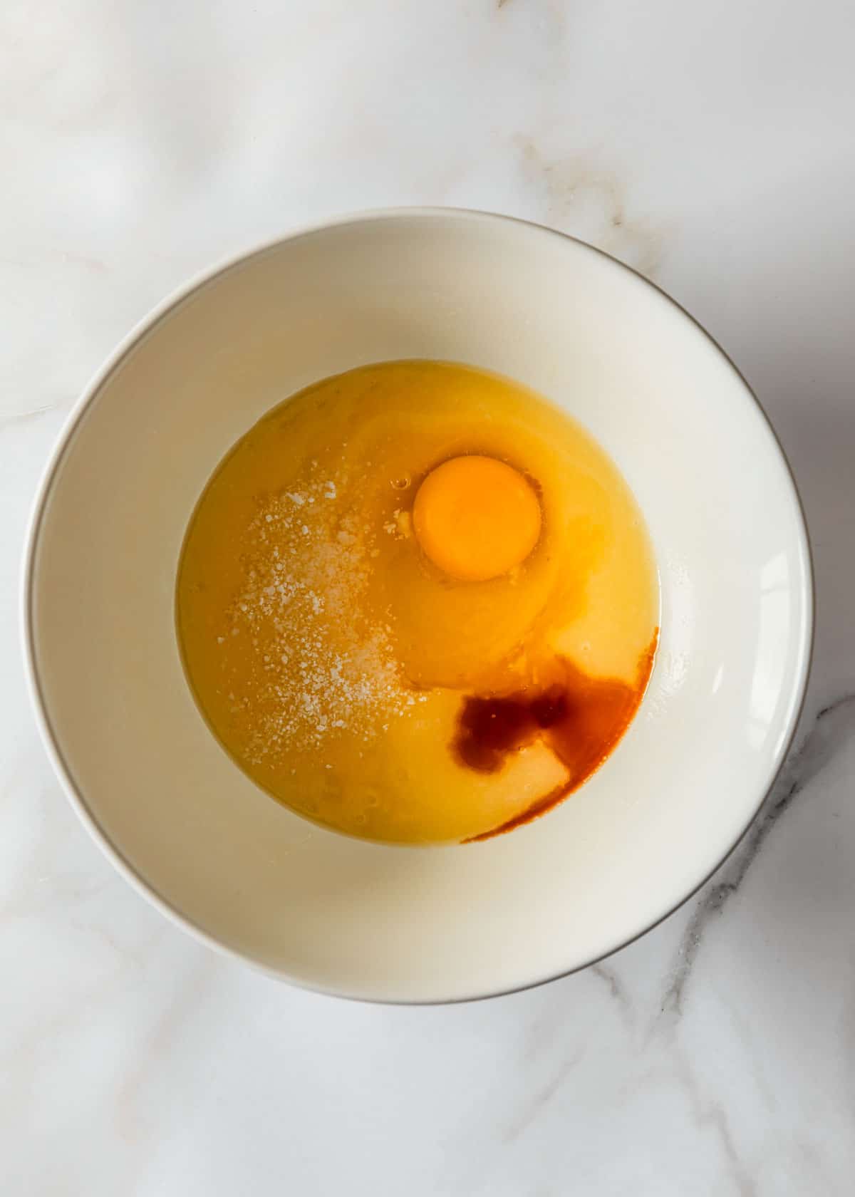 A white bowl of butter, sugar, an egg, and vanilla on a marble background.