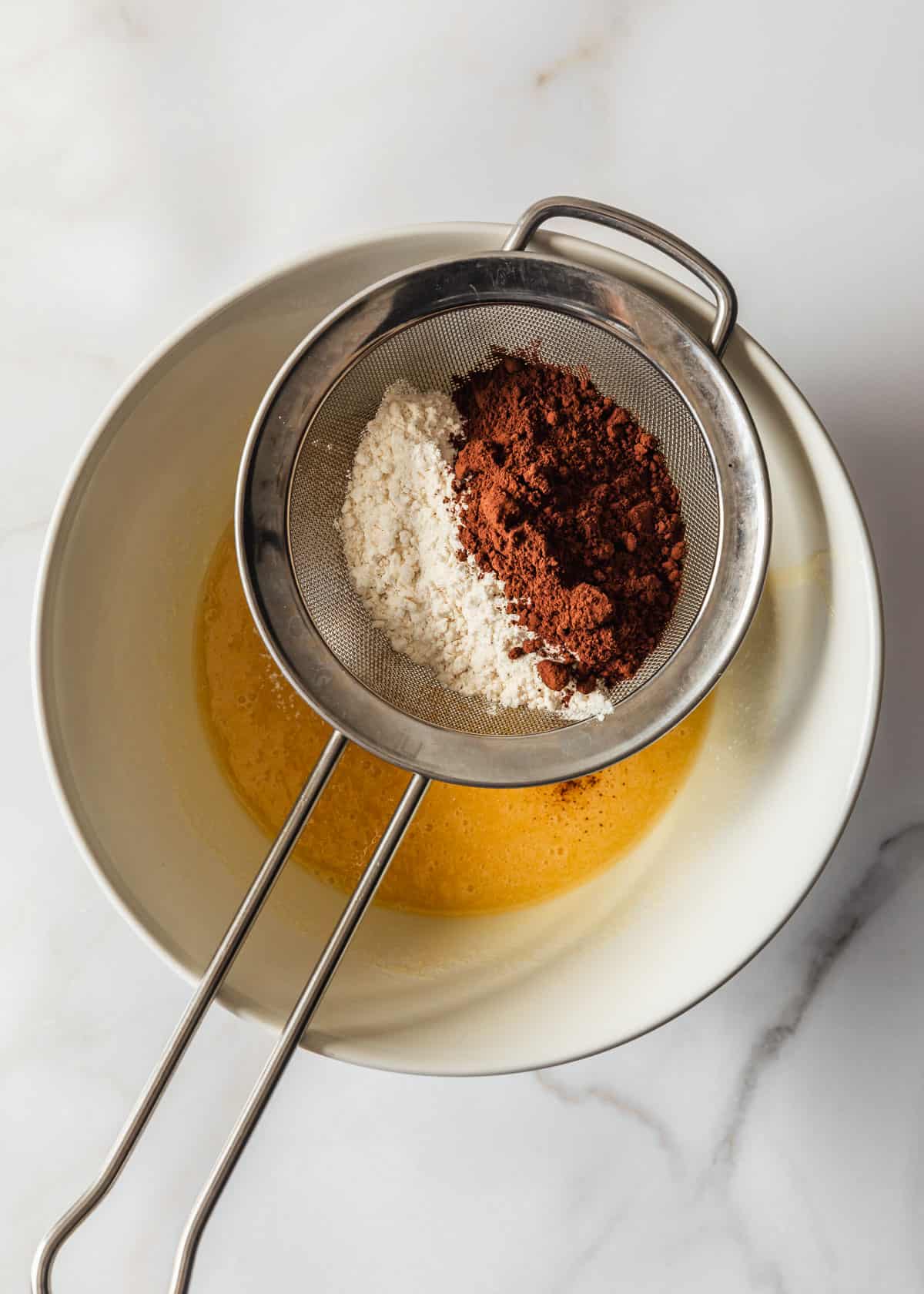 A white bowl of sugar and butter with a sifter of flour and cocoa powder on top.