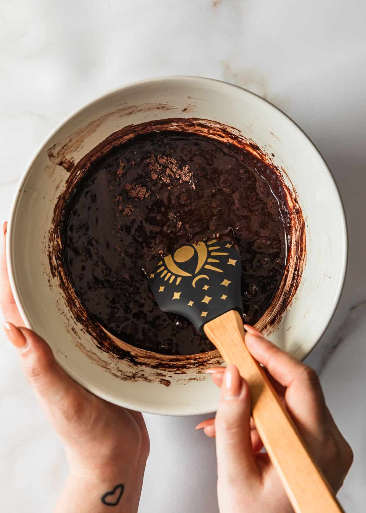 Hands using a rubber spatula to mix small batch brownies batter in a white bowl on a white counter.