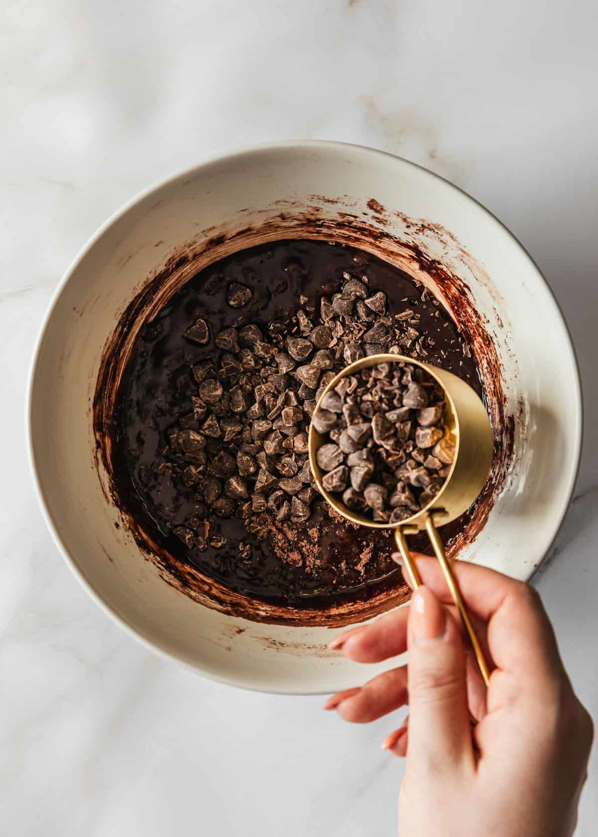 A hand pouring a measuring cup of chocolate chips into a white bowl of small batch brownies batter on a marble background.