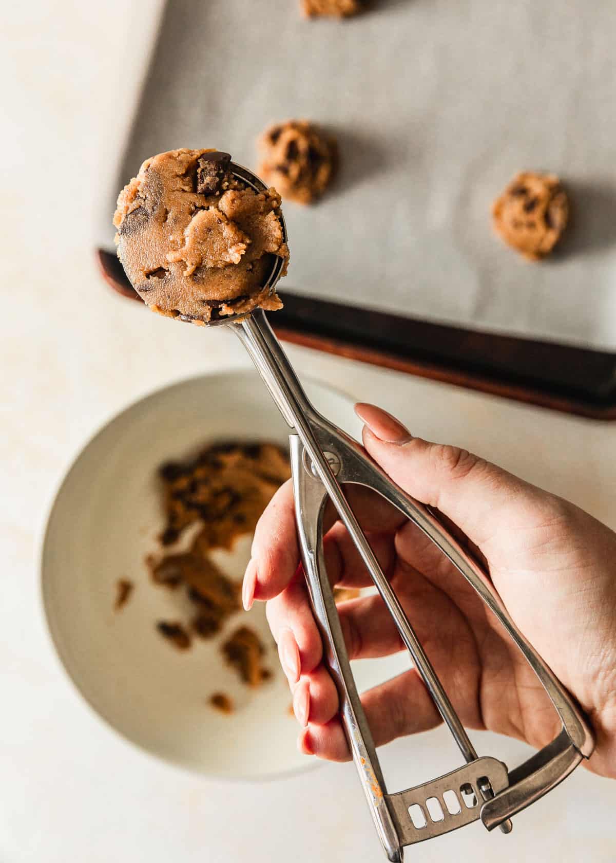 A hand holding a scoop of dough over a white bowl of small batch chocolate chip cookies dough.