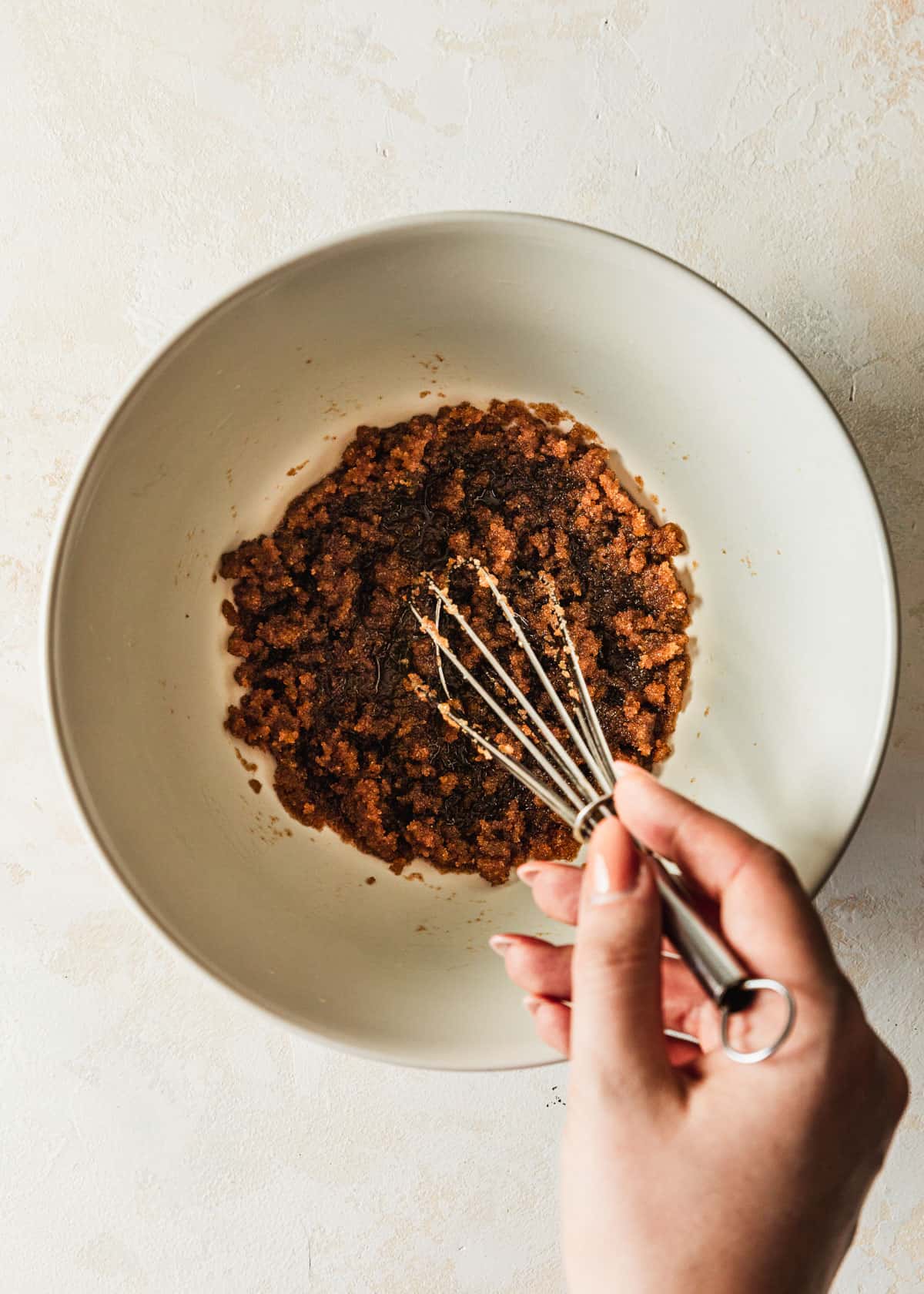A hand whisking butter and sugar in a white bowl on a tan background.