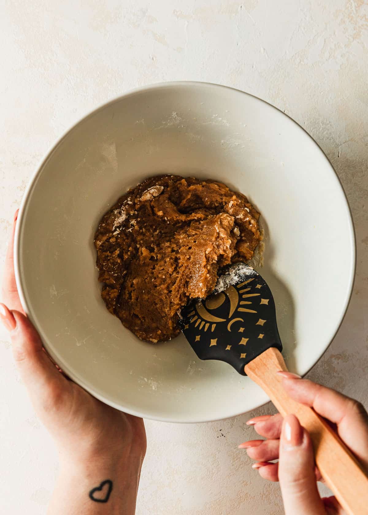A hand using a rubber spatula to mix a white bowl of cookie dough.
