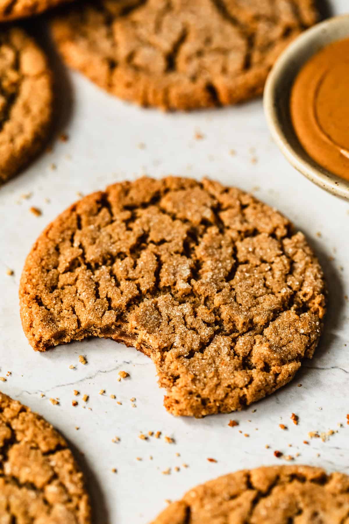 A cookie with a bite taken out of it next to a white bowl of peanut butter and rows of small batch peanut butter cookies with a white background.