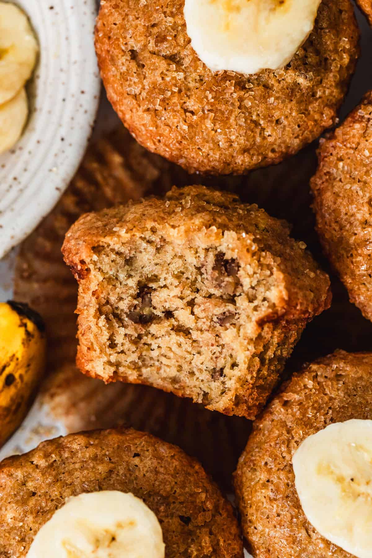 A muffin with a bite taken out of it next to a white bowl of banana slices and rows of small batch banana muffins.