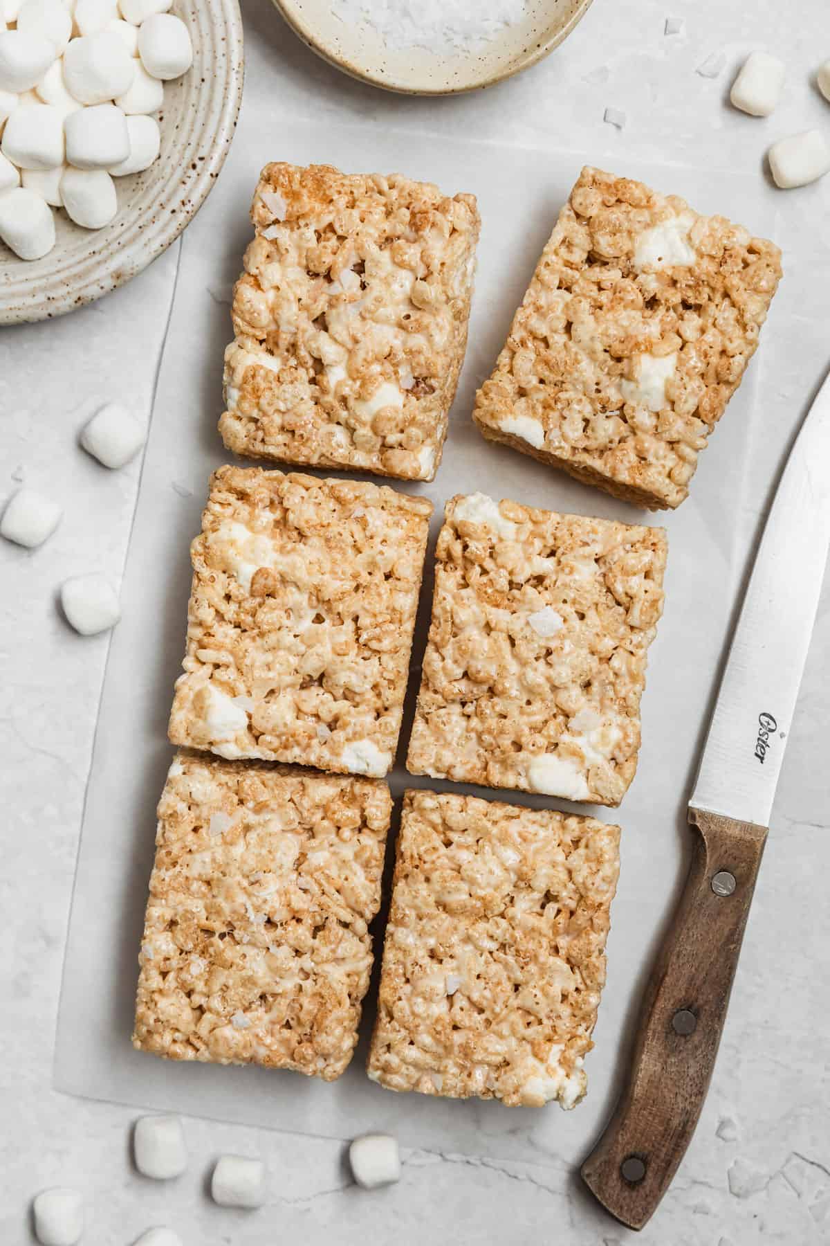 Rows of small batch Rice Krispies treats on a white counter next to a wood knife, white bowl of marshmallows, and white bowl of salt.
