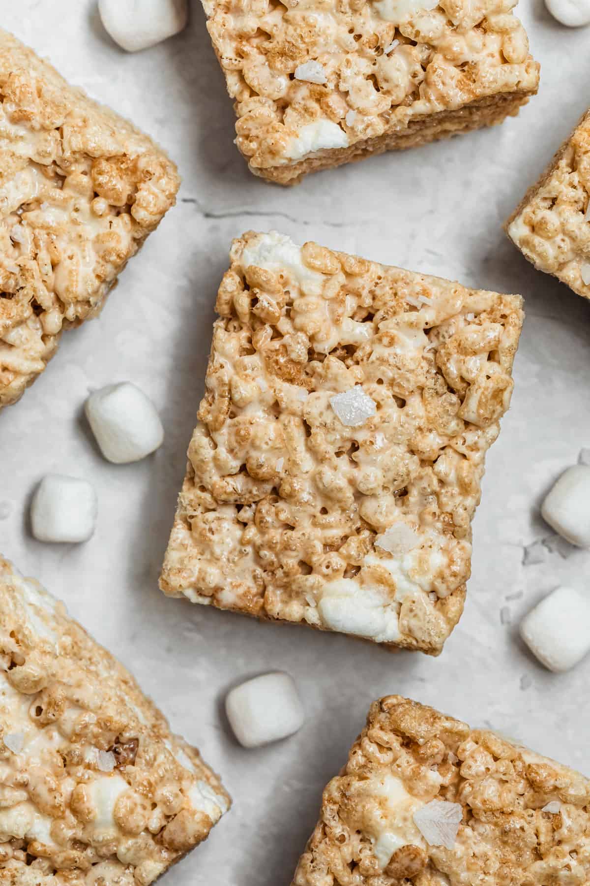 Rows of small batch Rice Krispies treats next to marshmallows on a white counter.