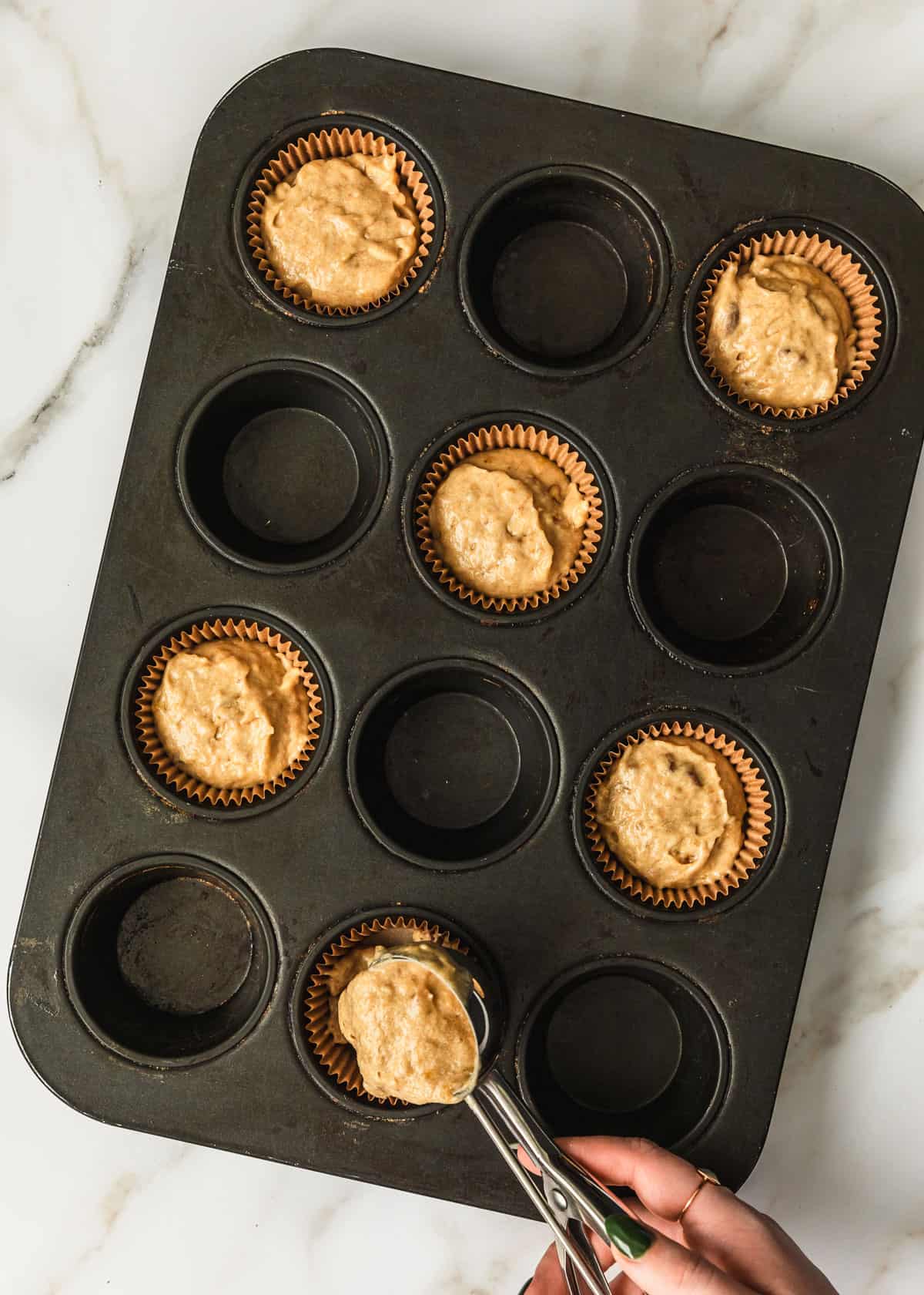 A hand using a scoop to add batter to muffin liners in a black pan.