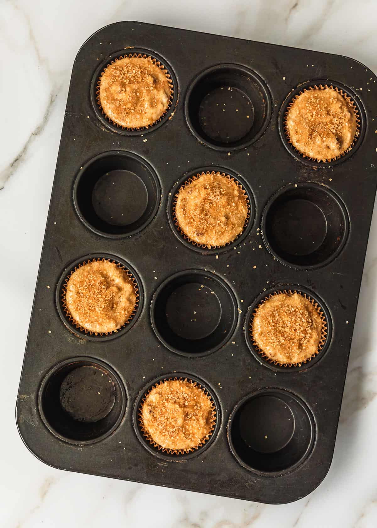 A black pan of one banana muffins batter with raw sugar on top.
