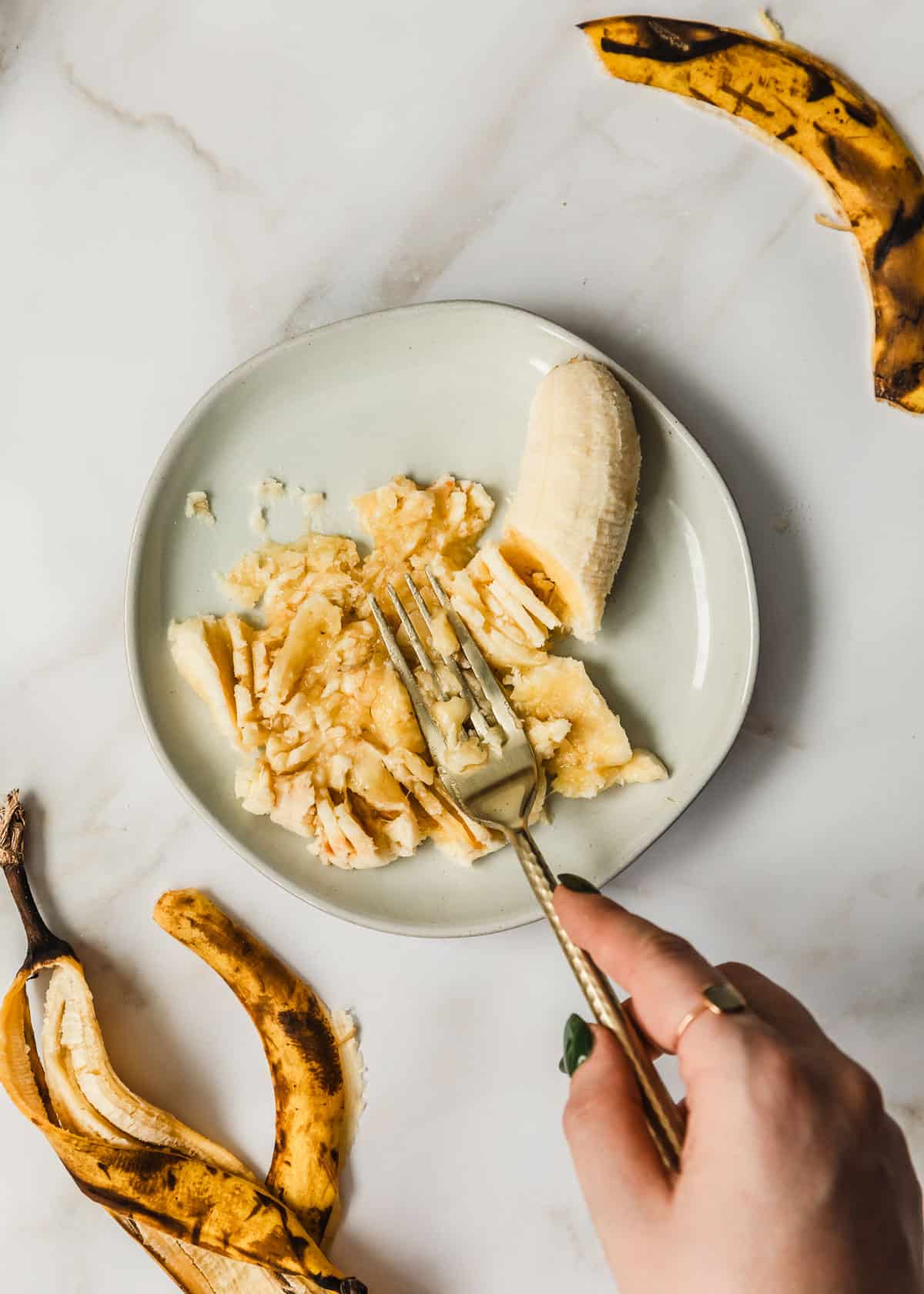 A hand mashing a banana on a white plate with a white background.