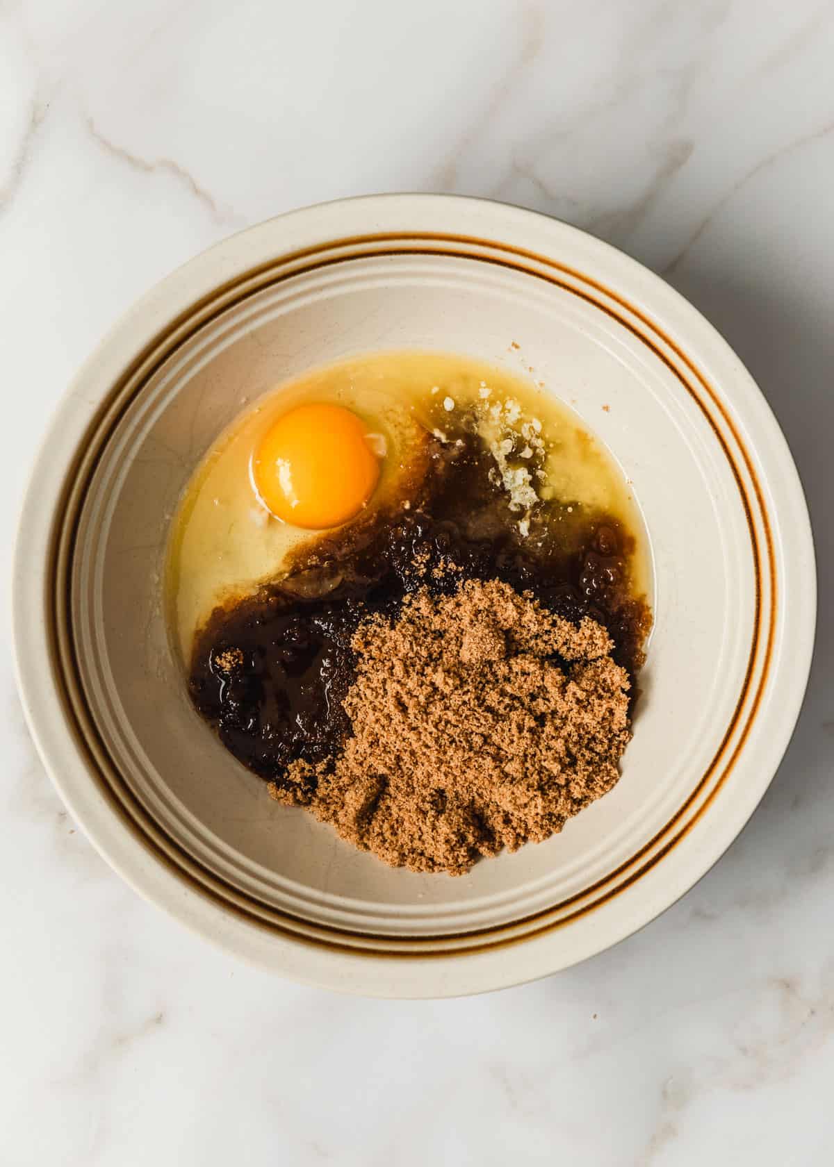 A white bowl of butter, brown sugar, an egg, and vanilla on a marble counter.