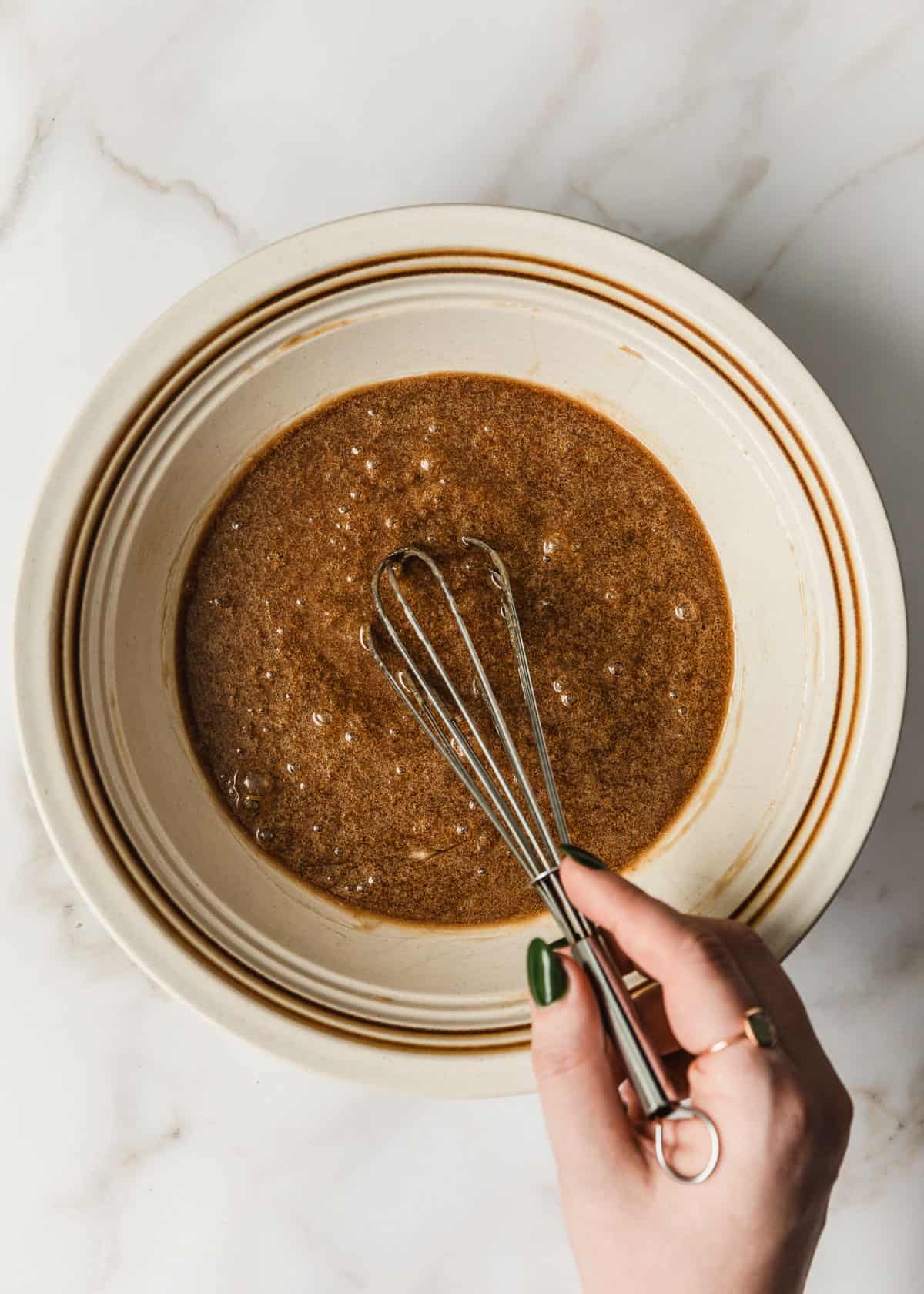 A hand whisking butter and brown sugar in a white bowl on a marble counter.