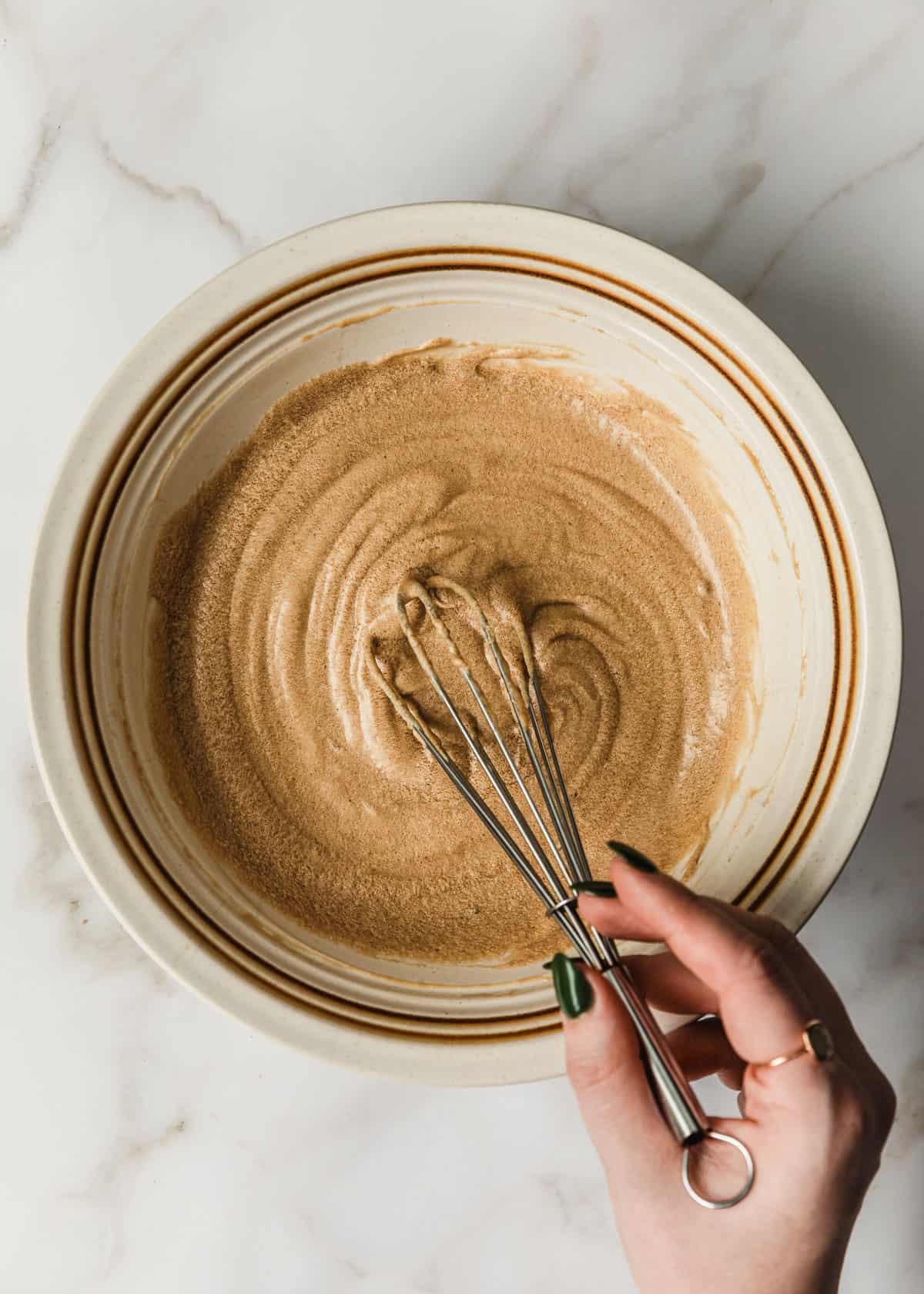 A hand using a whisk to mix butter, sugar, and an egg in a white bowl on a white counter.