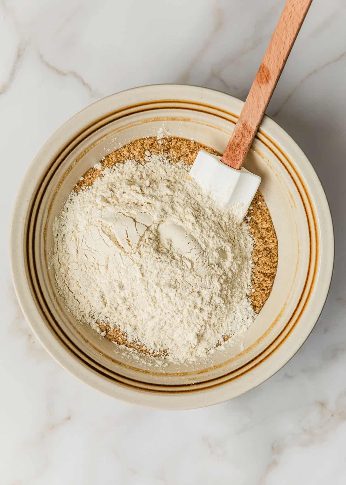 A white bowl with butter, sugar, banana, eggs, and flour on a marble table.