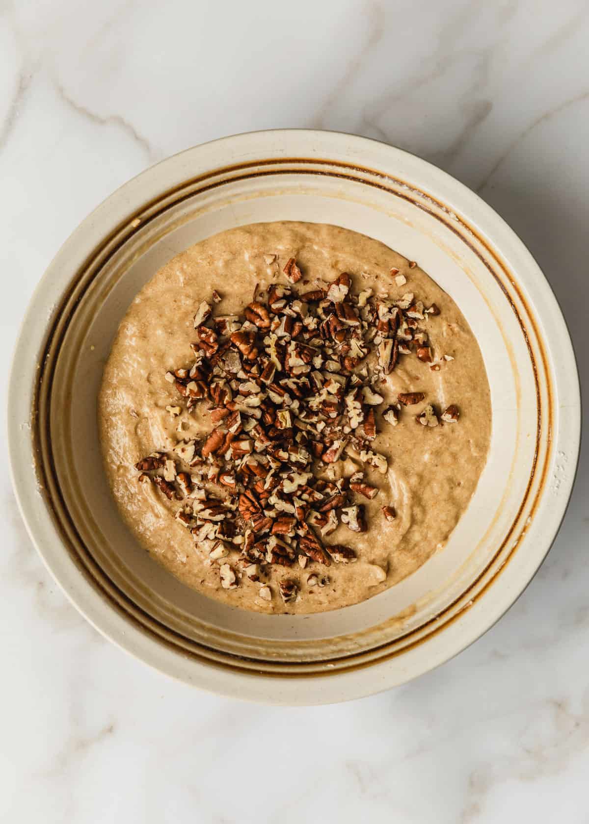 A white bowl of banana muffin batter and pecans on a white counter.