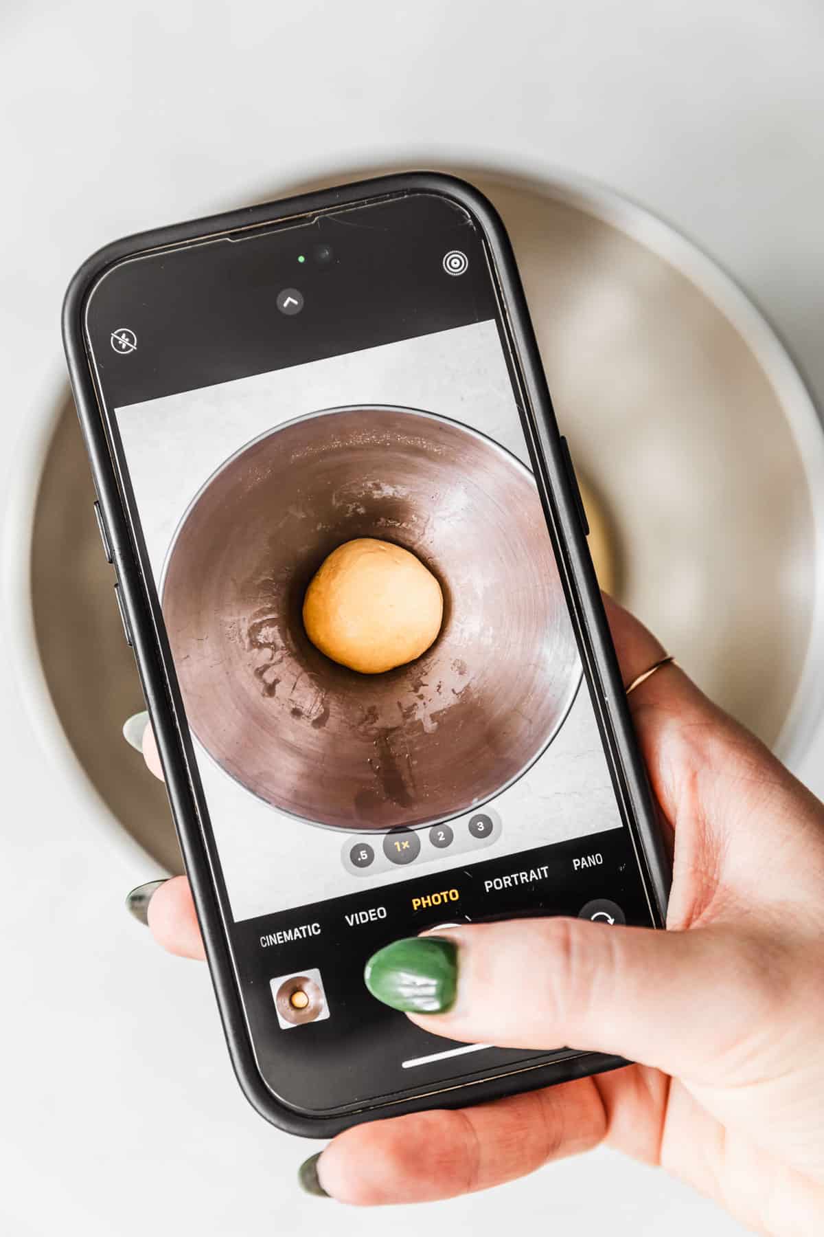 A hand using a phone to take a picture of a bowl of dough.