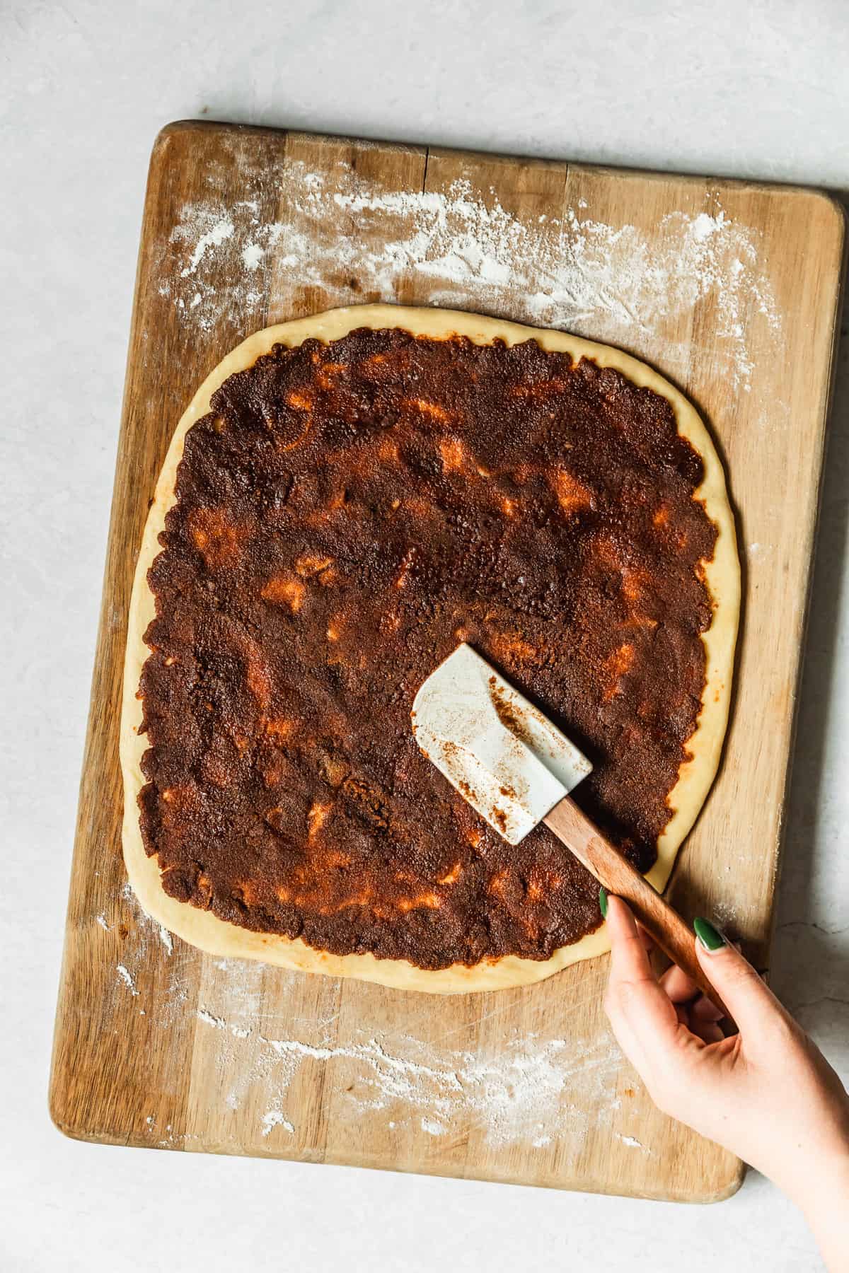 A hand using a rubber spatula to spread cinnamon sugar on dough.