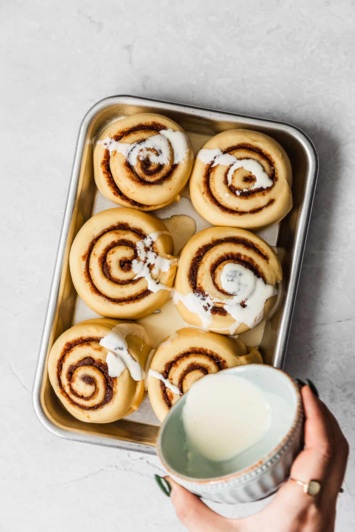 A hand pouring cream over a pan of raw small batch cinnamon rolls.