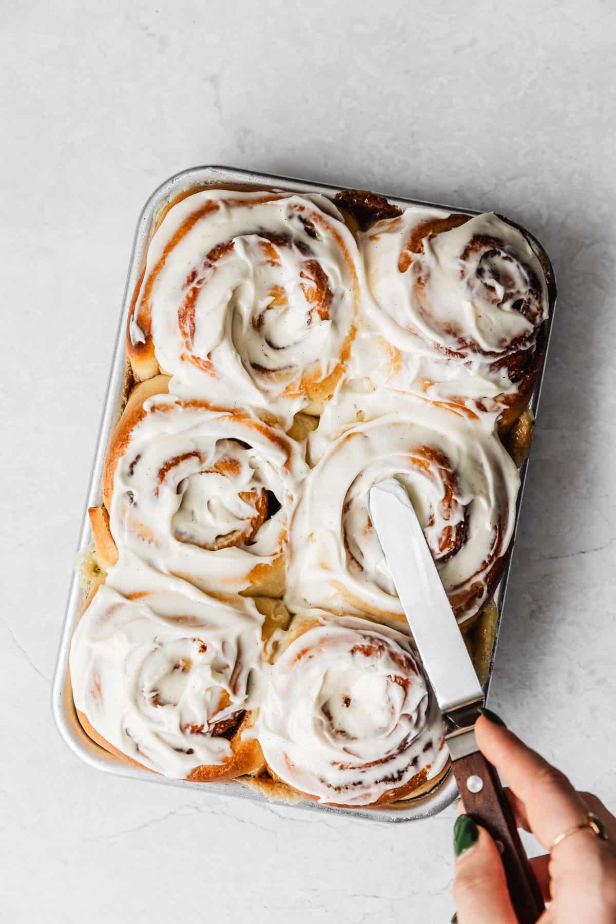 Hands using an offset spatula to frost a pan of small batch cinnamon rolls on a white counter.