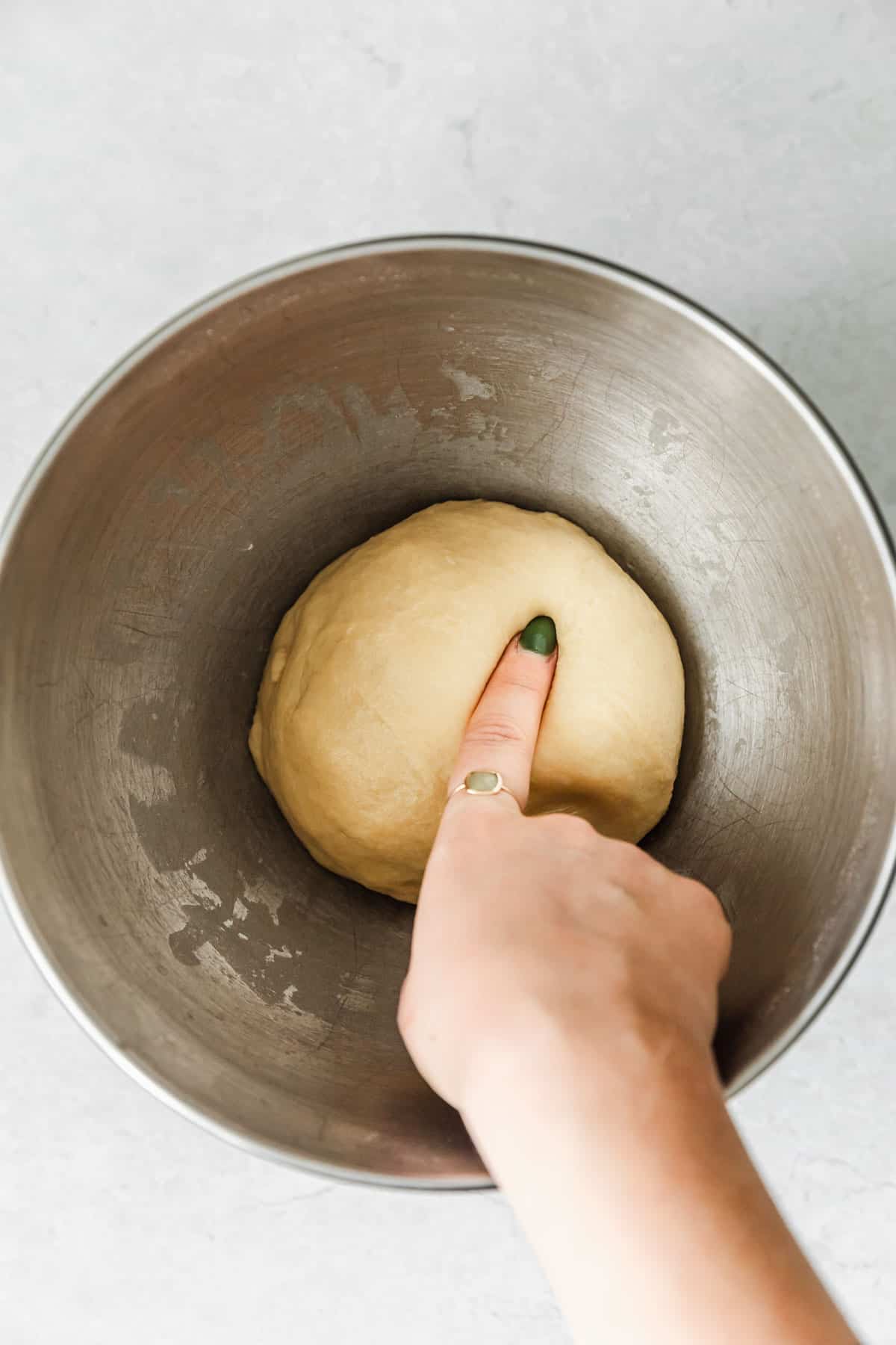 A hand poking dough in a silver bowl to do the poke test.