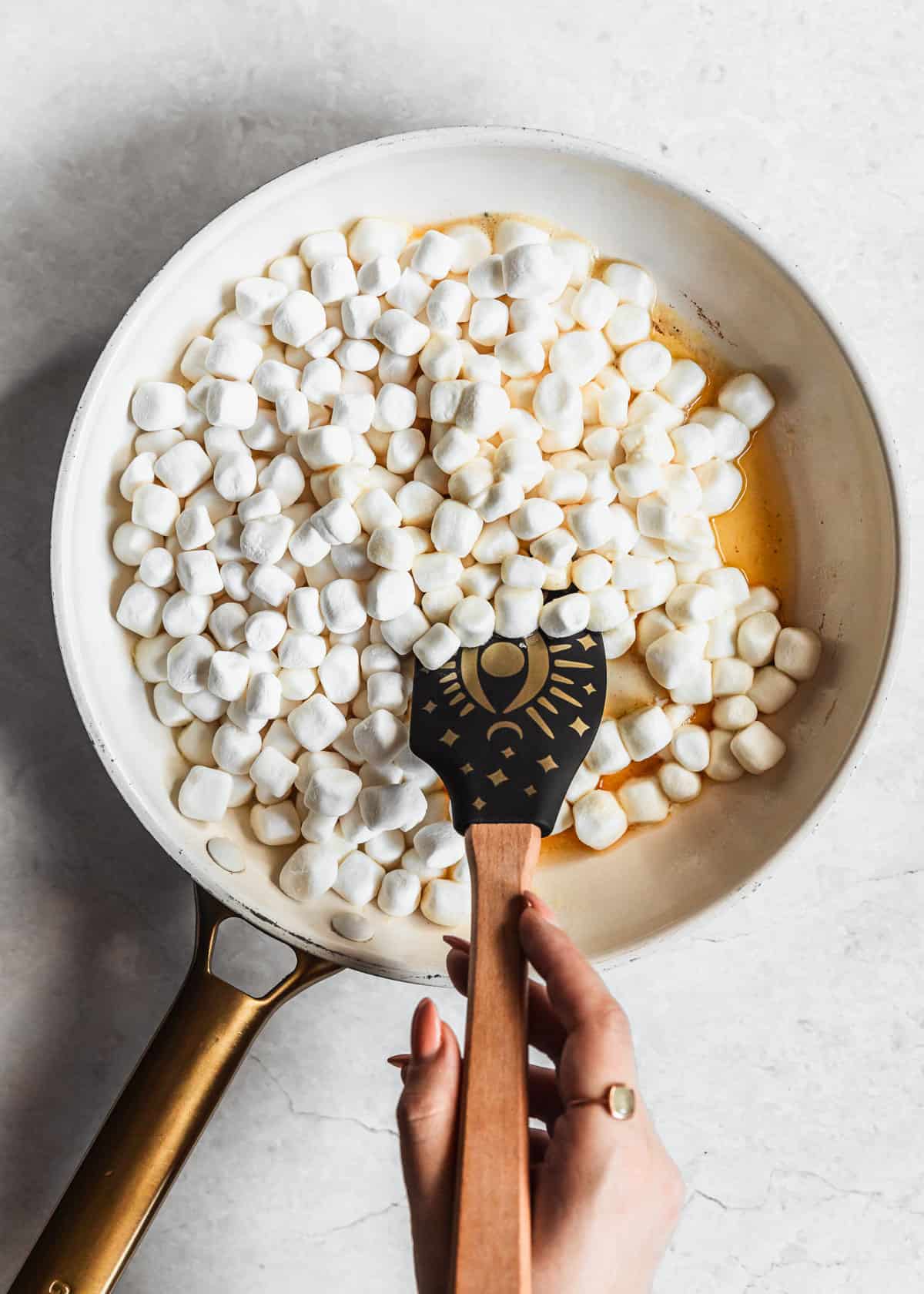 A hand using a rubber spatula to stir marshmallows in a white pan on a marble counter.