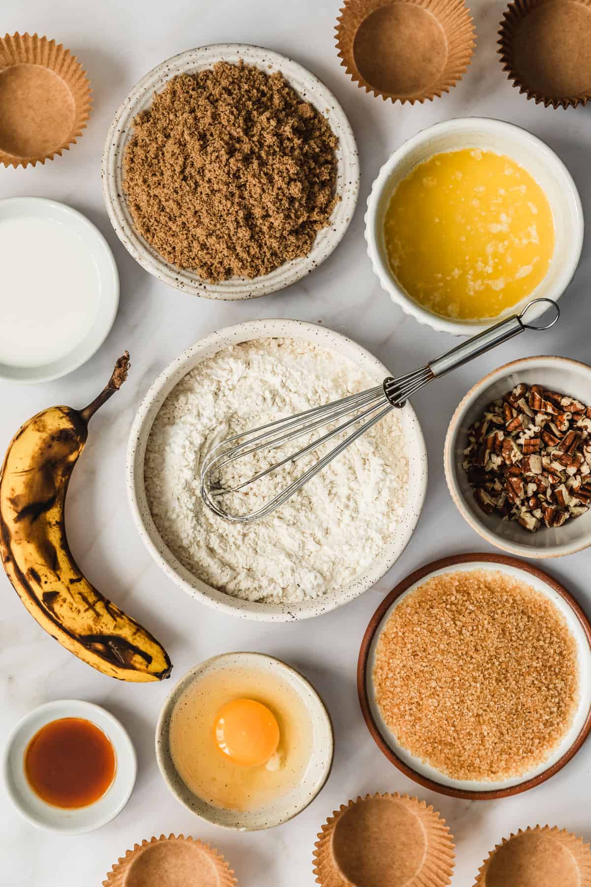 White and brown bowls of flour, brown sugar, melted butter, milk, an egg, vanilla, nuts, and raw sugar next to a banana on a white counter.