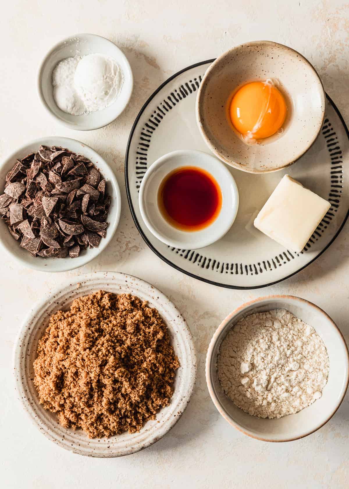 White bowls of brown sugar, flour, chocolate, vanilla, butter, an egg yolk, cornstarch, salt, and baking soda on a tan counter.