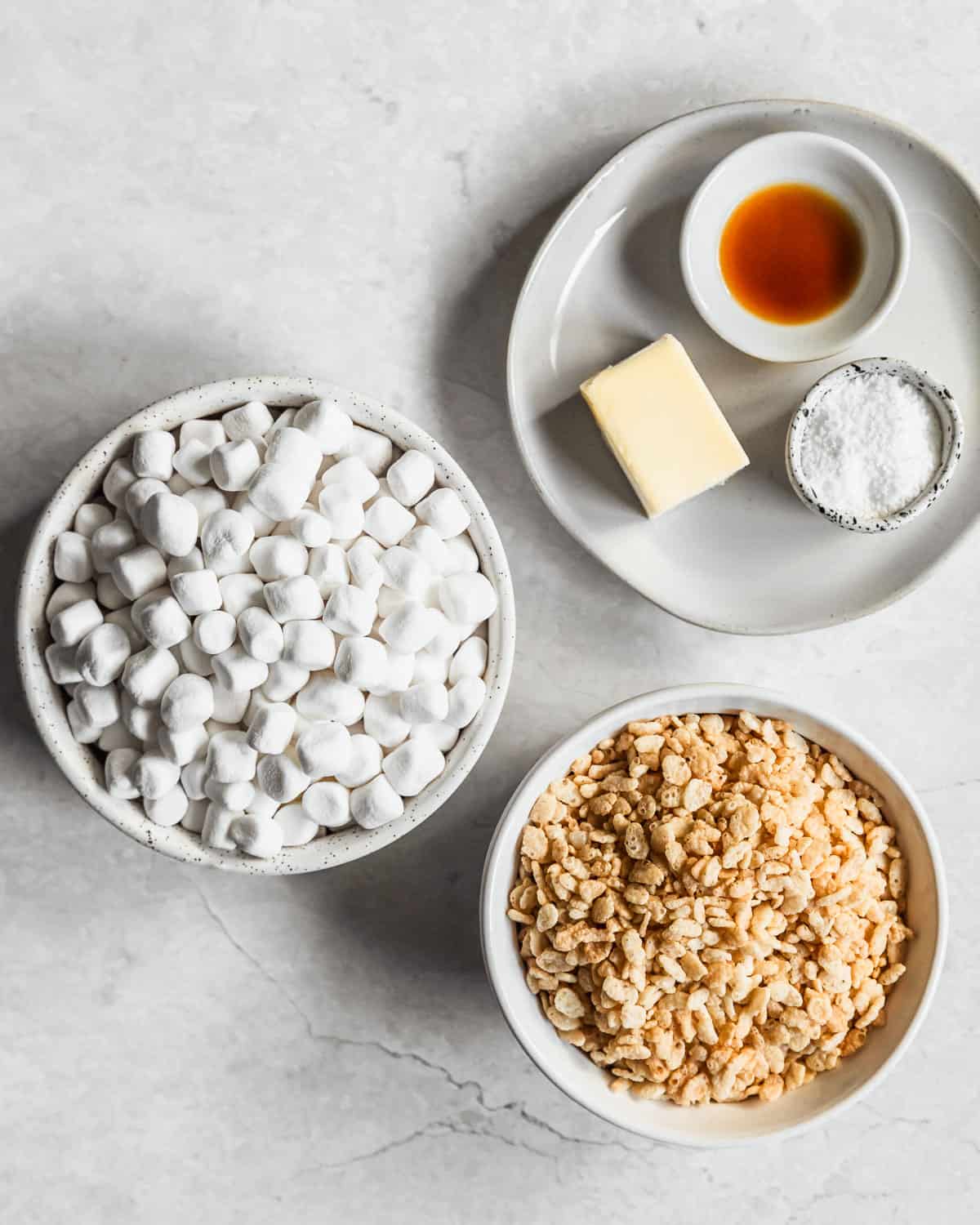 White bowls of marshmallows, cereal, vanilla, butter, and salt on a white counter.