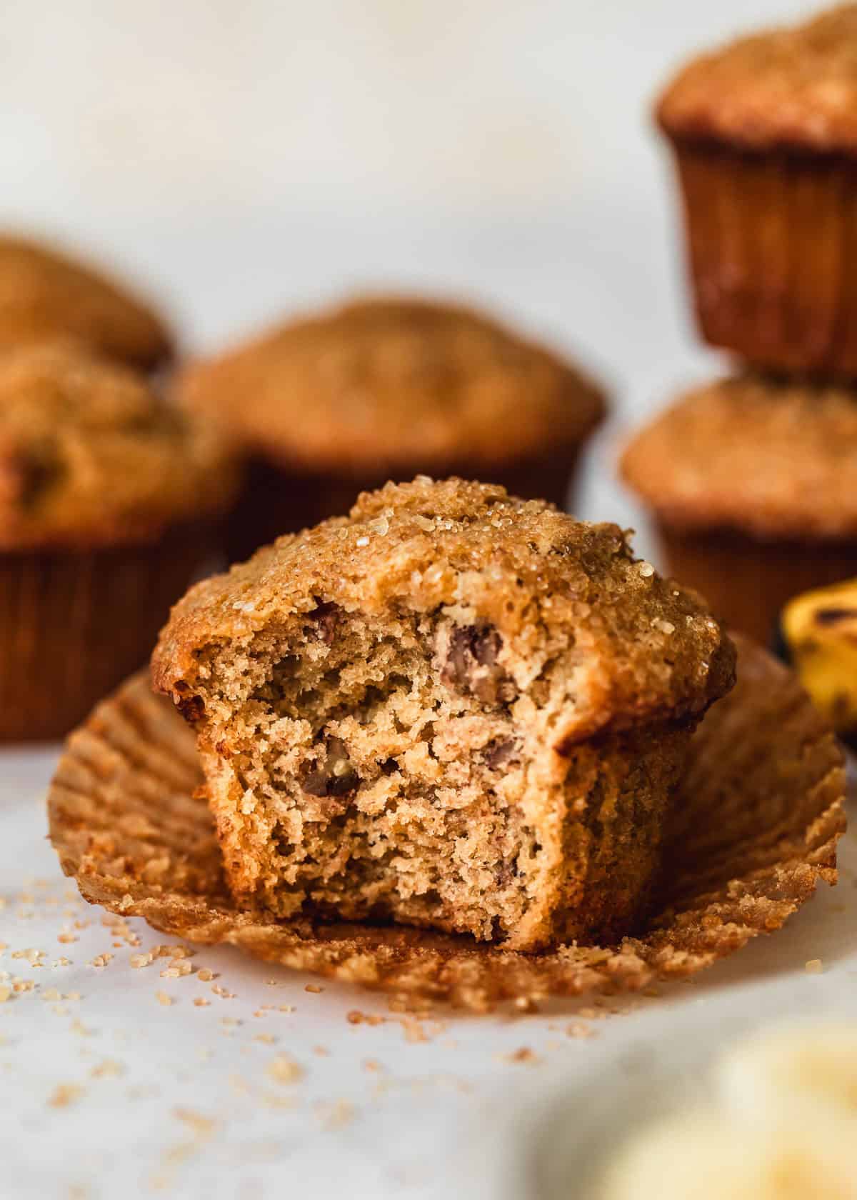 A muffin with a bite taken out of it on a white counter next to rows of small batch banana muffins and a white bowl of banana slices.