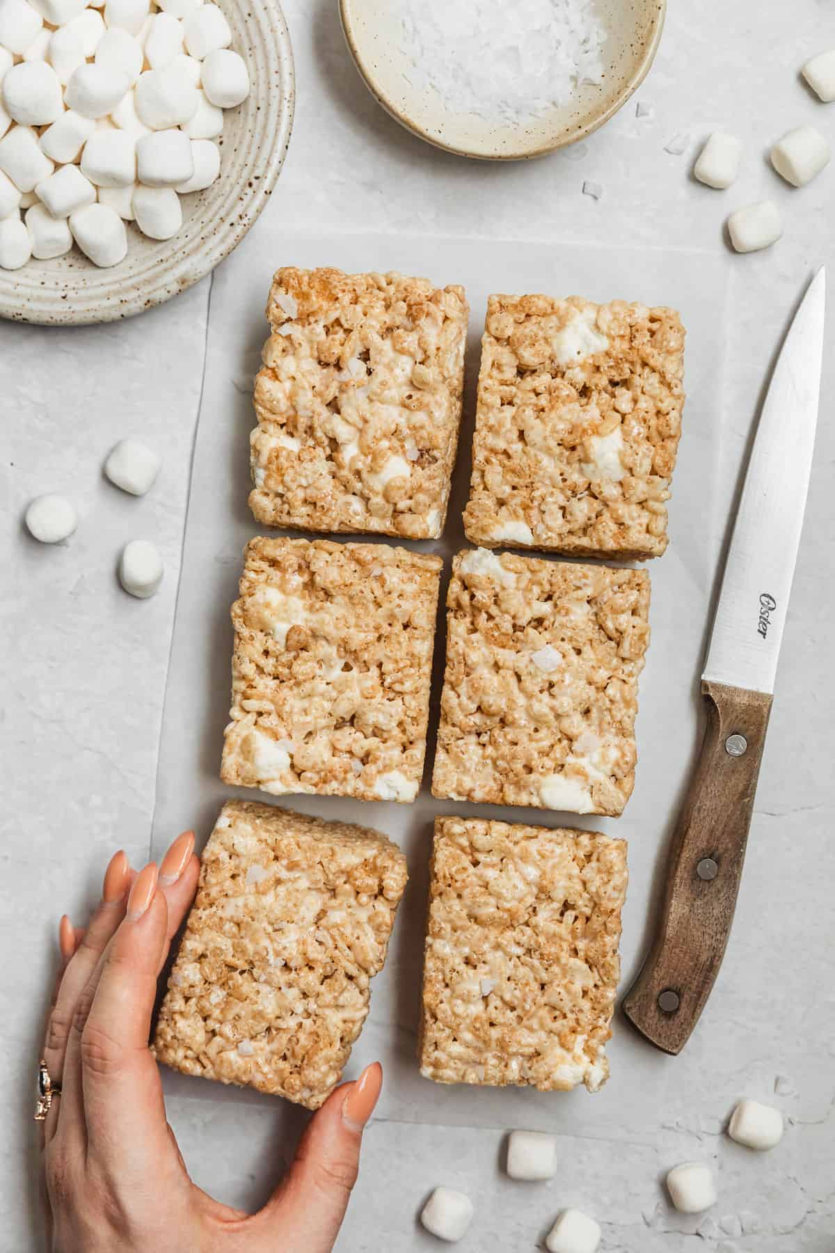 A hand grabbing a bar from a batch of small batch Rice Krispies treats on a white counter next to a wood knife and white bowls of marshmallows and flaky salt.