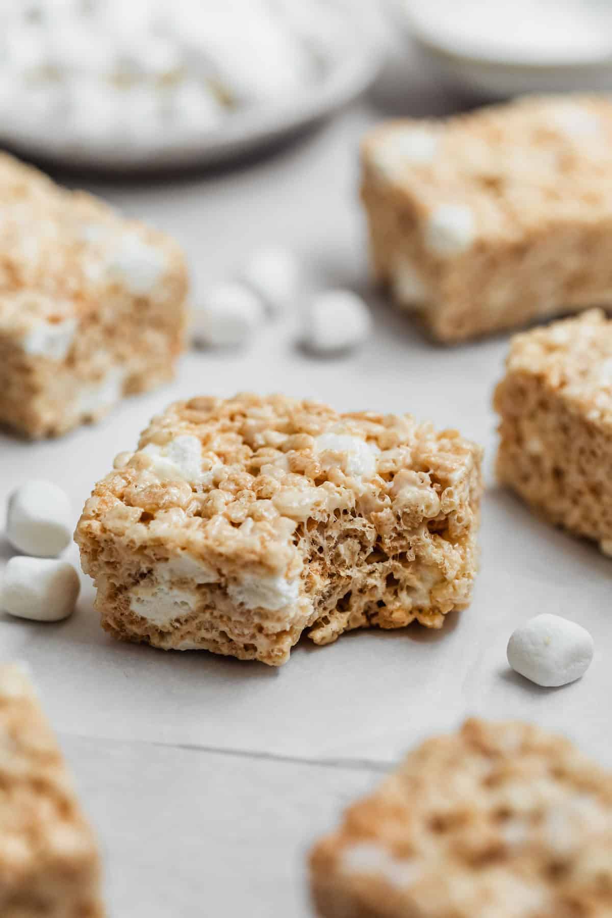 A cereal treat with a bite taken out of it on a white counter next to rows of small batch Rice Krispies treats and a white bowl of marshmallows.