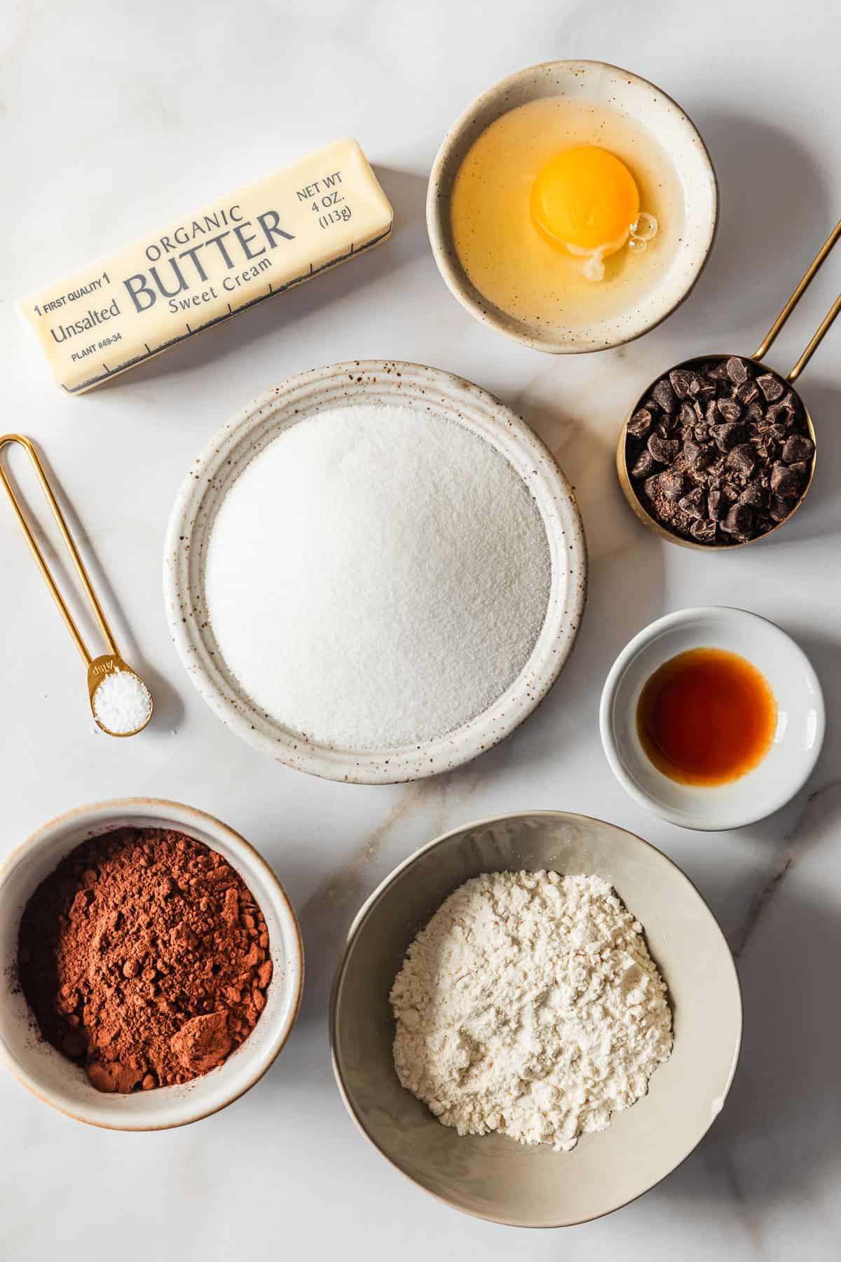 White bowls of sugar, cocoa powder, flour, vanilla, and an egg on a white counter next to a stick of butter, a teaspoon of salt, and a measuring cup with chocolate chips.