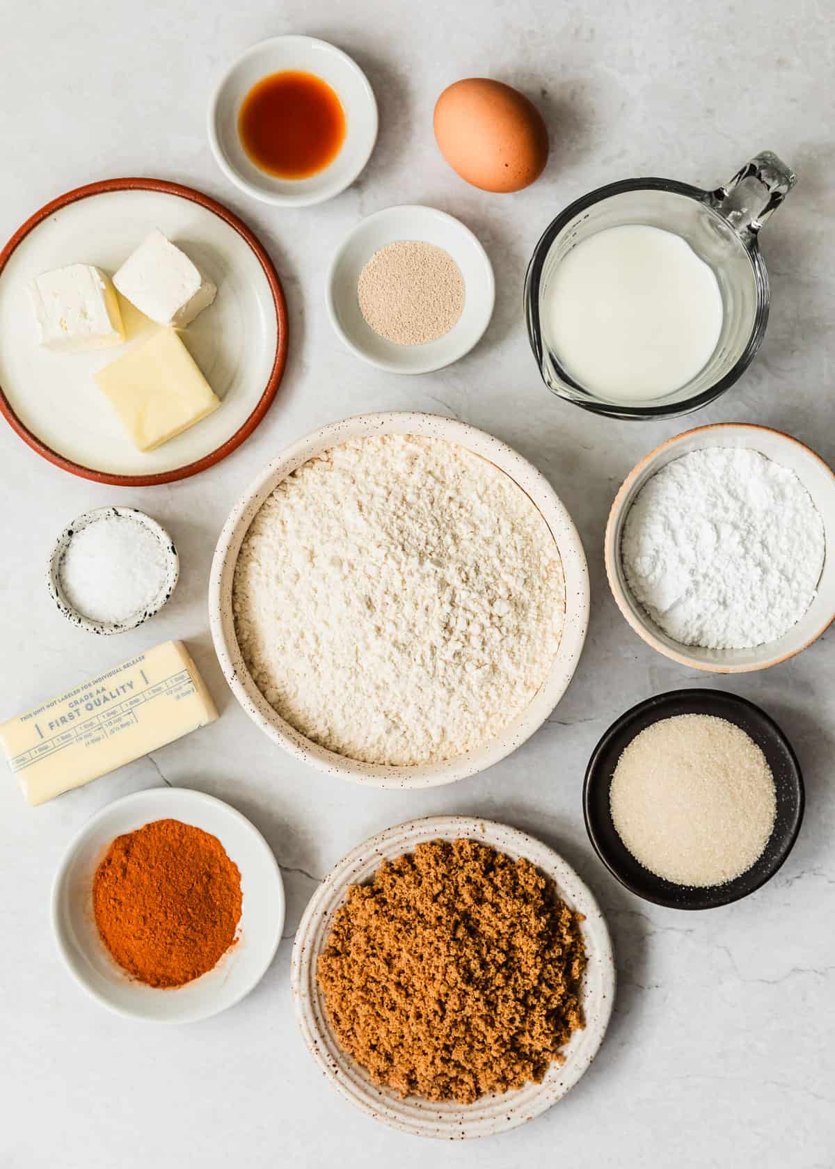White bowls of flour, brown sugar, cinnamon, sugar, powdered sugar, salt, butter, yeast, and milk on a white counter.