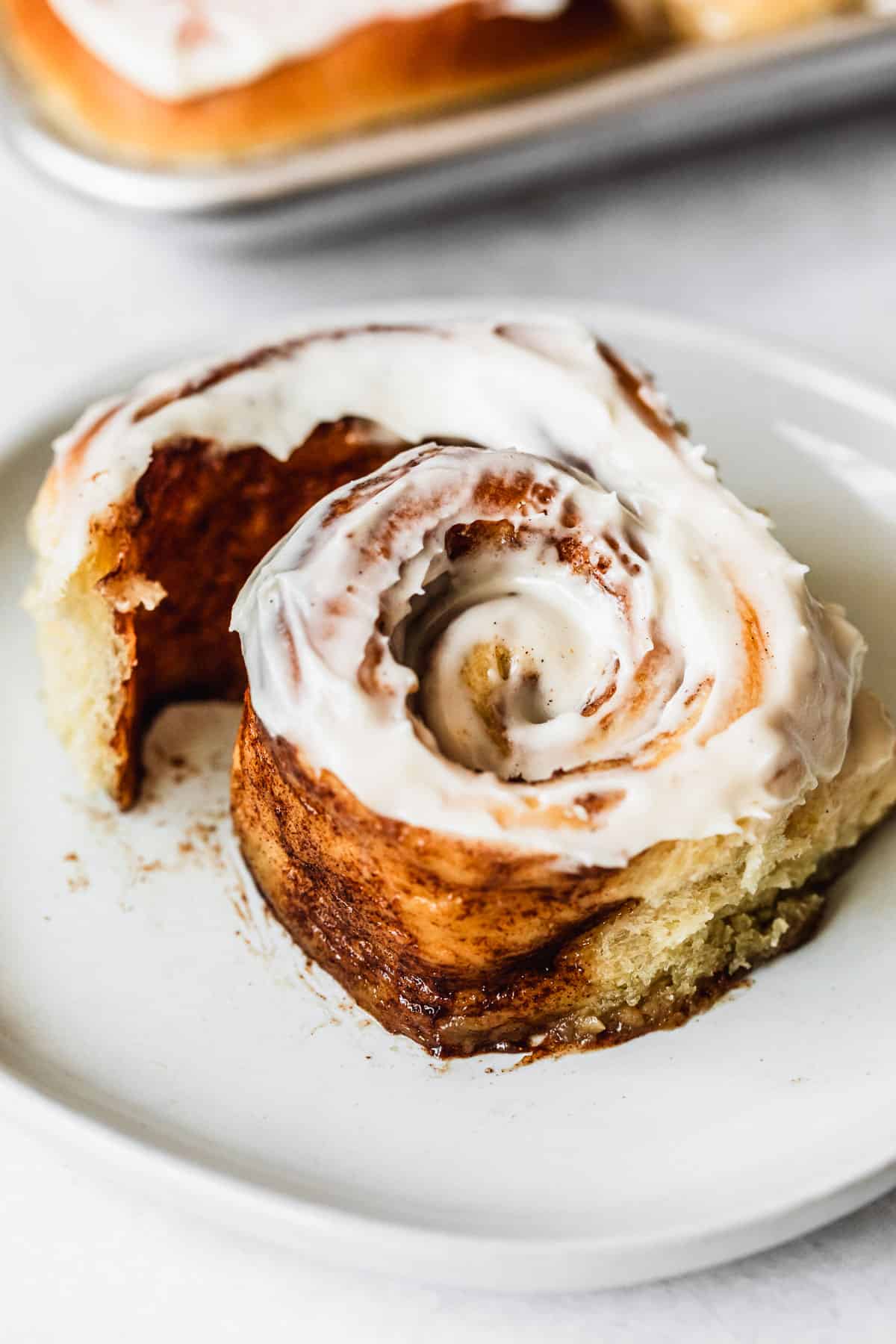 A cinnamon roll unraveling on a white plate next to a pan of small batch cinnamon rolls with a white background.