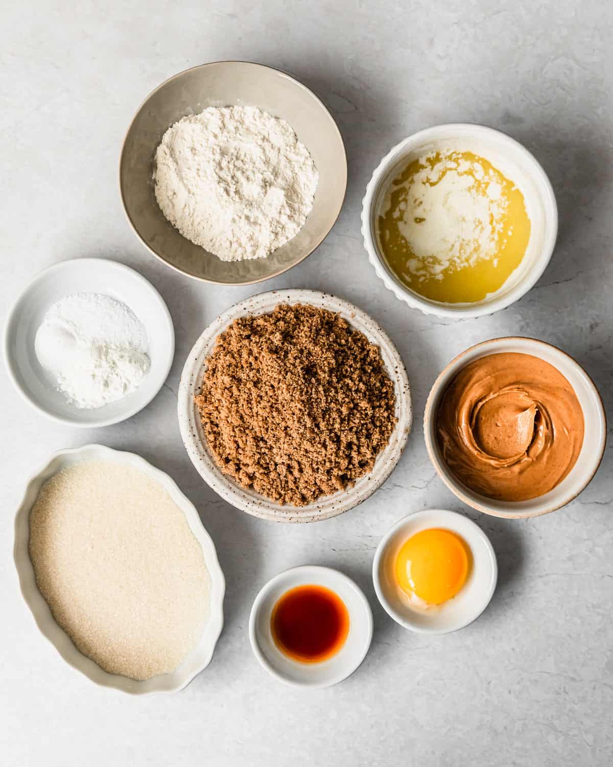 White bowls of melted butter, flour, baking soda, salt, brown sugar, peanut butter, white sugar, vanilla, and an egg yolk on a white counter.