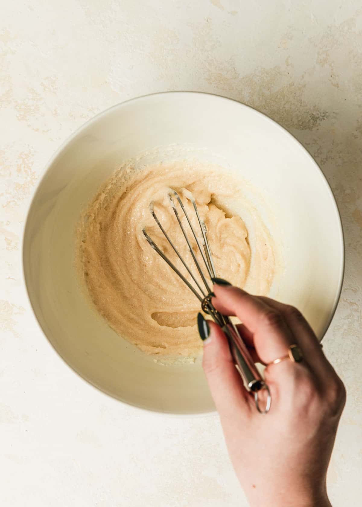 A hand whisking brown butter and sugar in a white bowl on a tan counter.