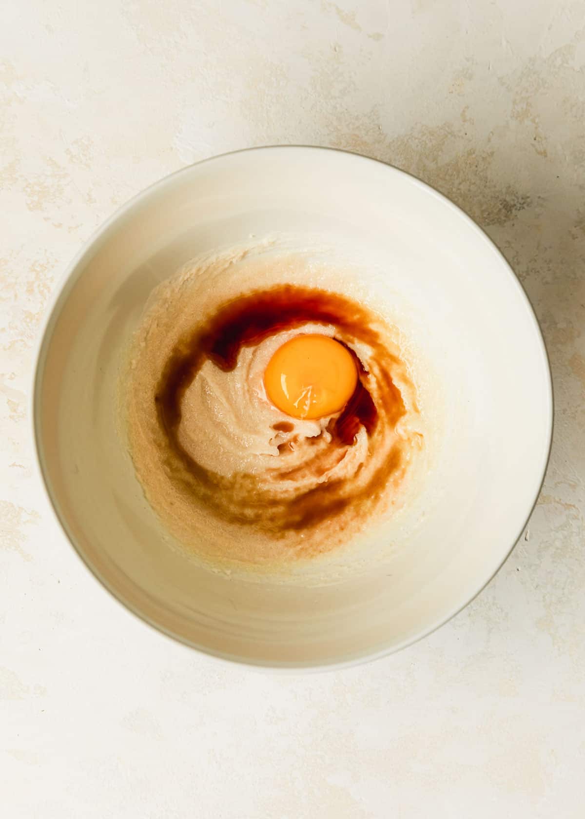 A white bowl of brown butter, sugar, an egg yolk, and vanilla on a beige table.