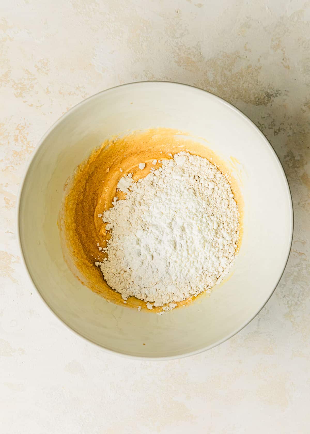 A white bowl of butter, sugar, eggs, vanilla, and flour on a beige table.