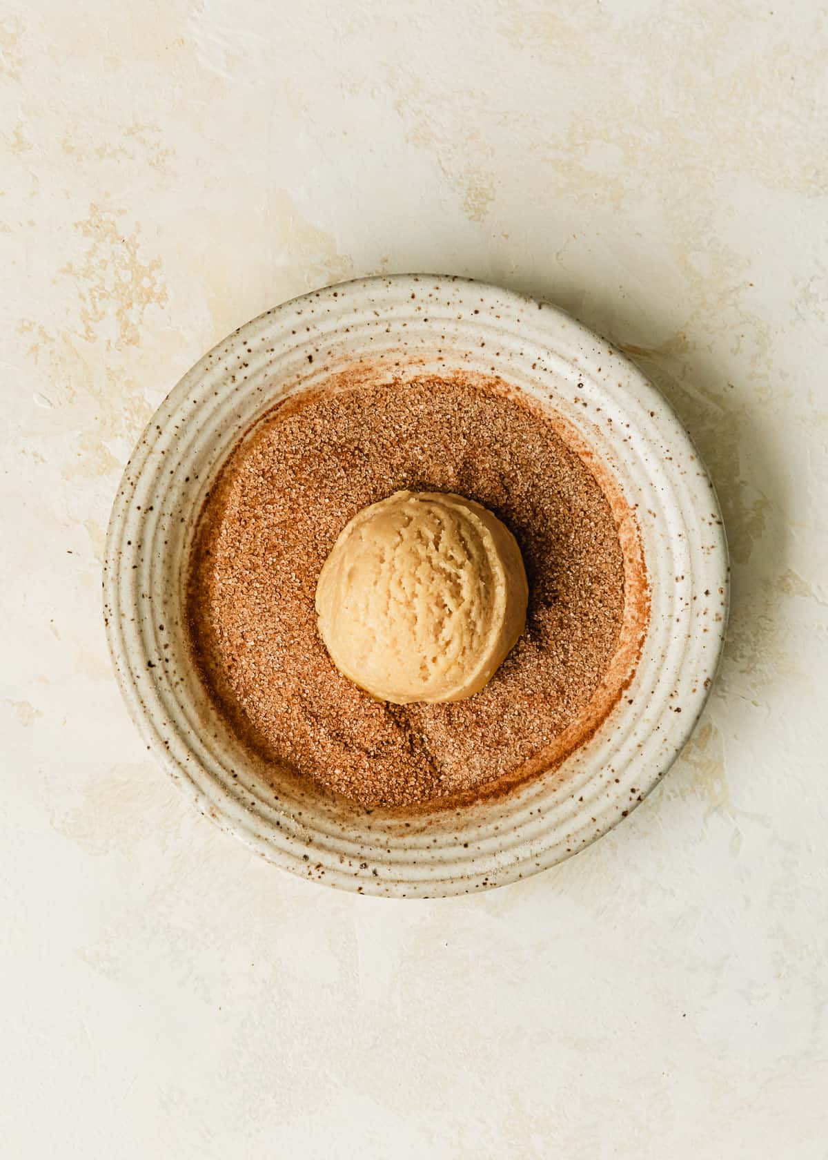 A white bowl of cinnamon sugar with a cookie dough bowl on a beige counter.