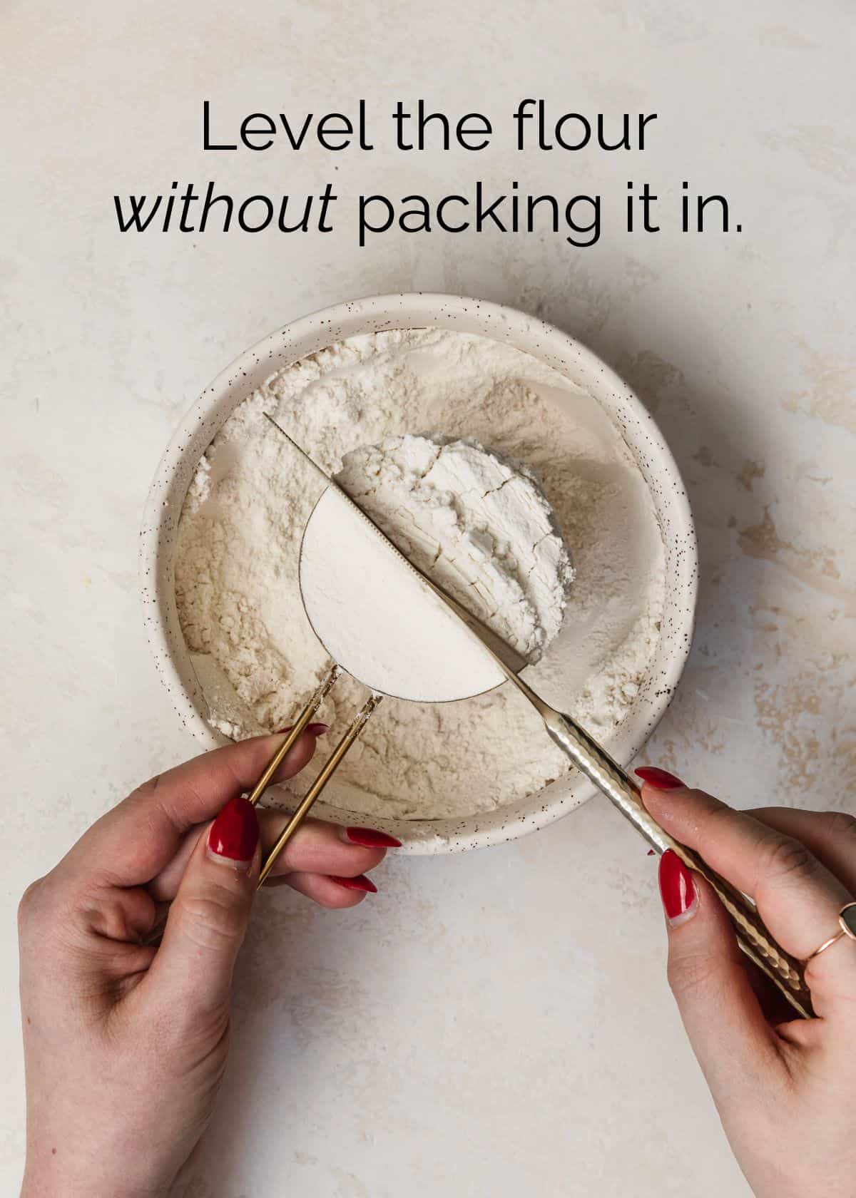 A hand using a knife to level flour in a measuring cup on a tan counter.