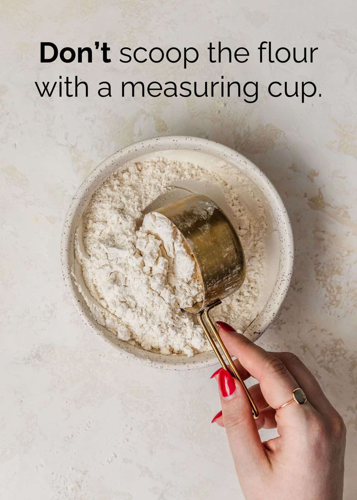 A hand using a gold measuring cup to scoop flour out of a white bowl on a tan counter.