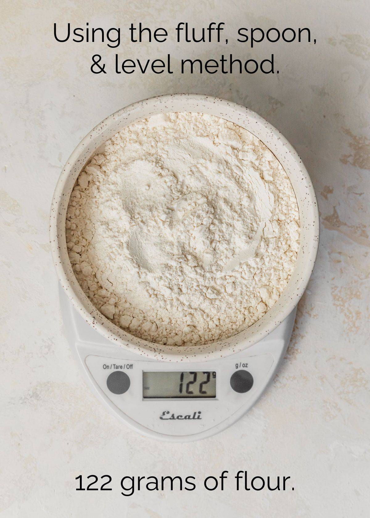 How to measure ingredients using the fluff, spoon, and level method. A white bowl of flour on a scale with a stone background.