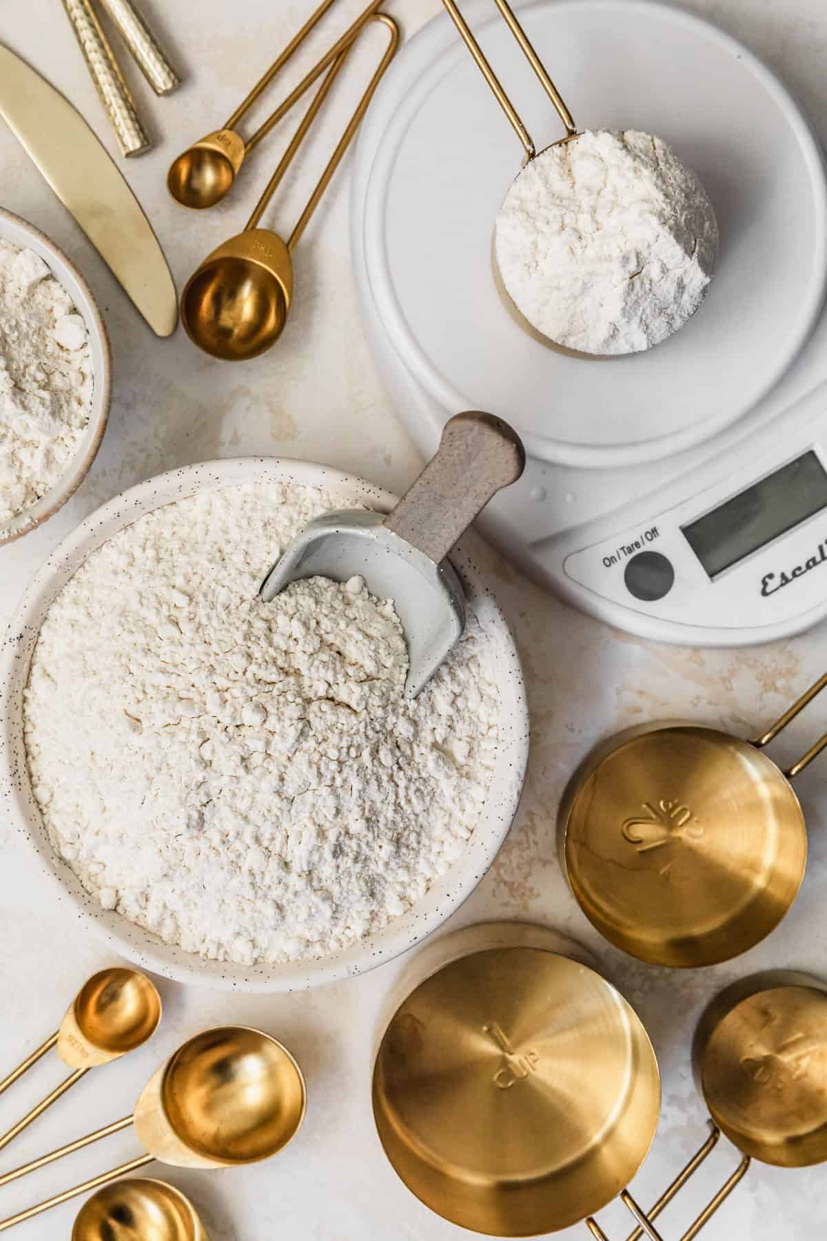 Gold measuring cups, white bowls of flour, and a kitchen scale on a tan counter.