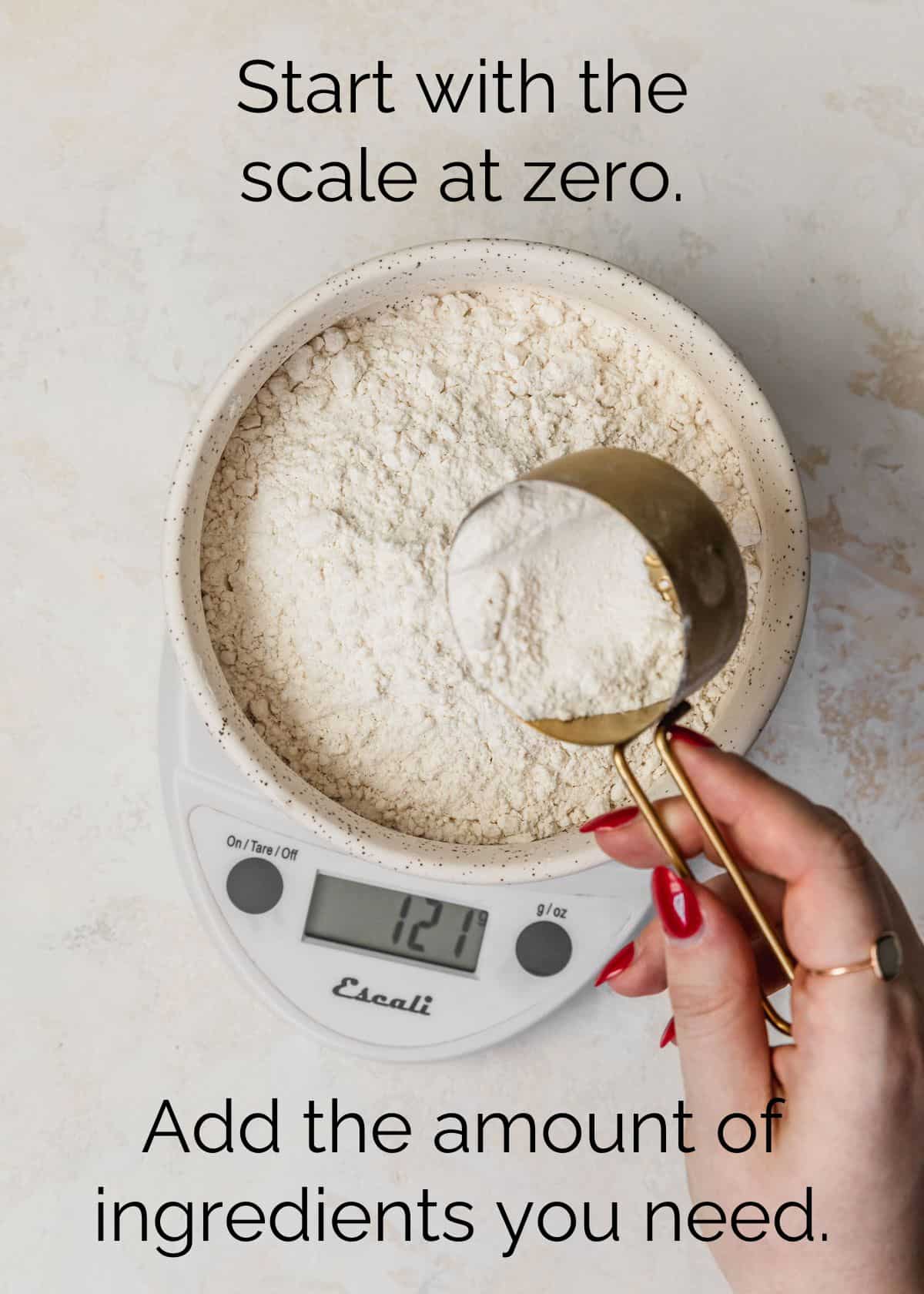 How to measure ingredients for small batch baking. A hand pouring flour from a gold measuring cup into a white bowl on a kitchen scale with a tan background.