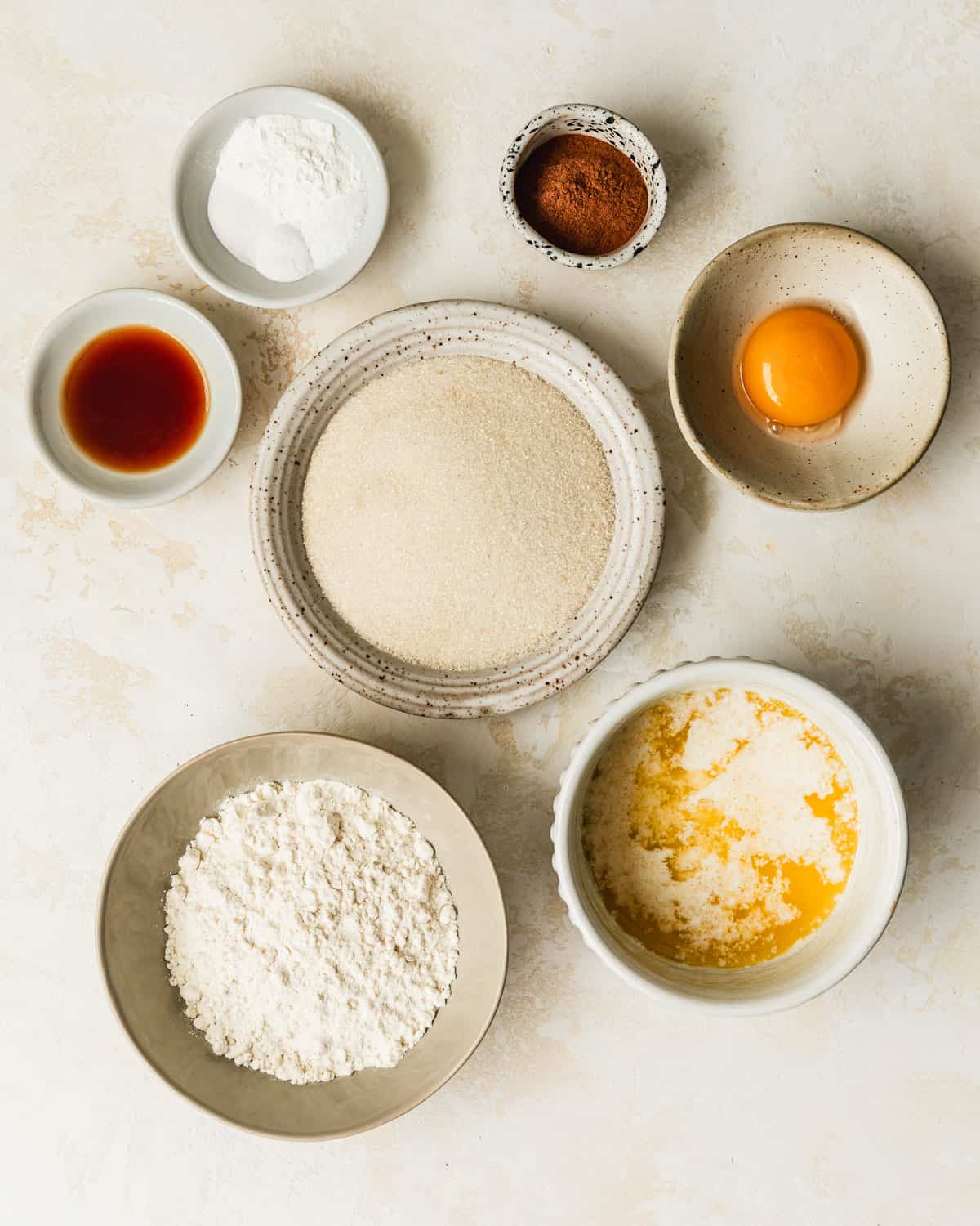 White bowls of sugar, flour, butter, an egg yolk, vanilla, cinnamon, salt, baking soda, and cream of tartar on a tan counter.