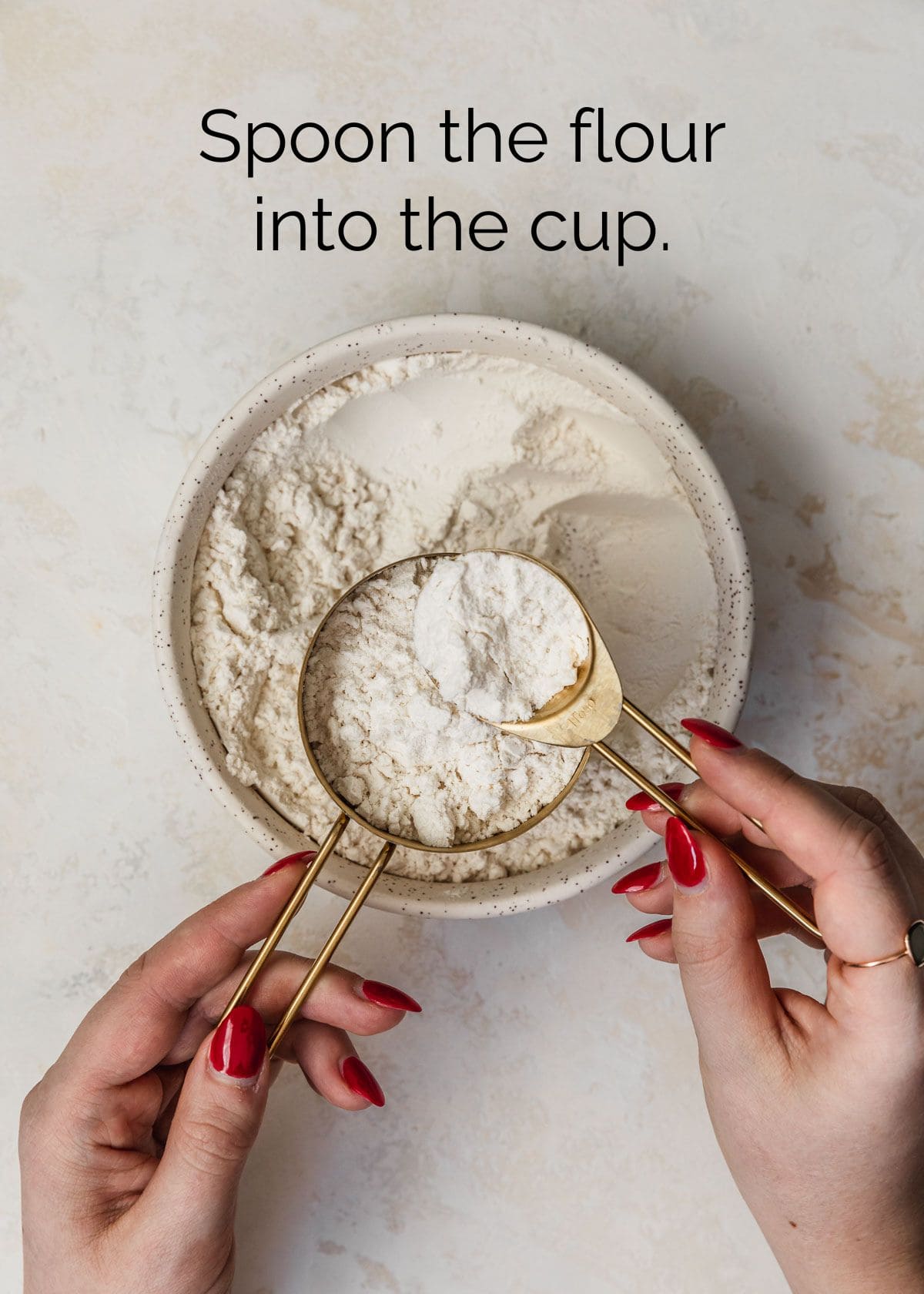 How to measure ingredients for small batch baking with measuring cups. A hand spooning flour into a measuring cup on a tan counter.