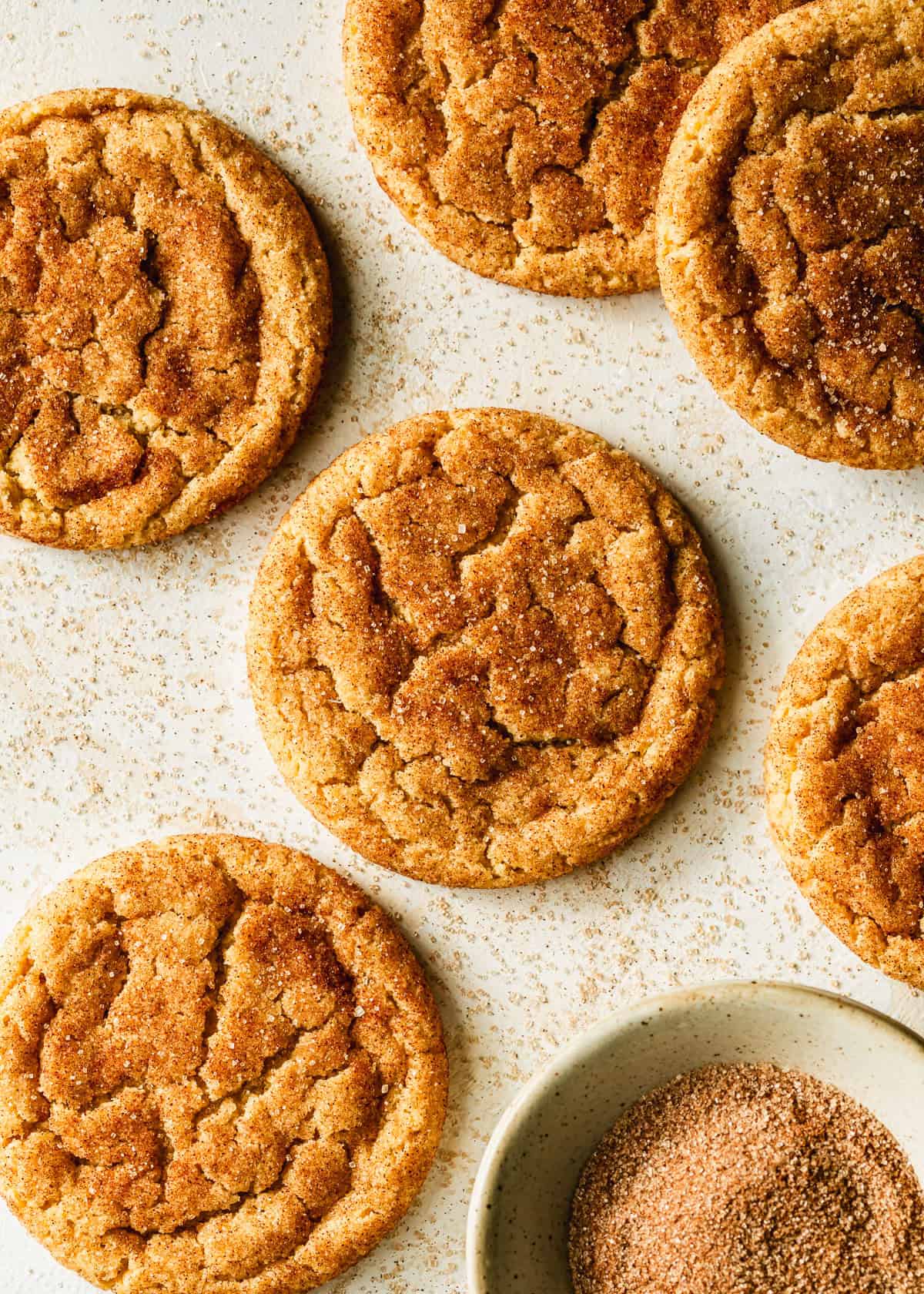 Rows of small batch snickerdoodles and a white bowl of cinnamon sugar on a tan stone counter.