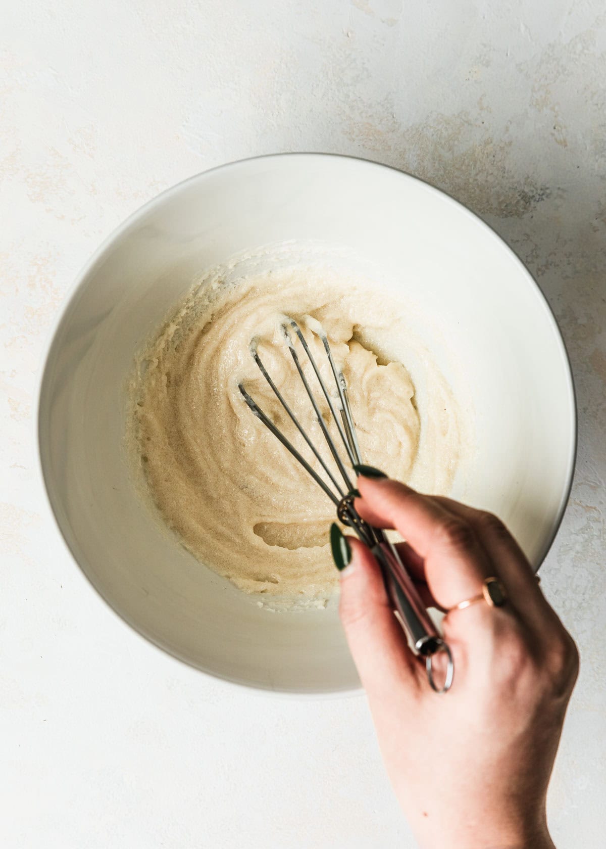 A hand whisking butter and sugar in a white bowl on a white counter.